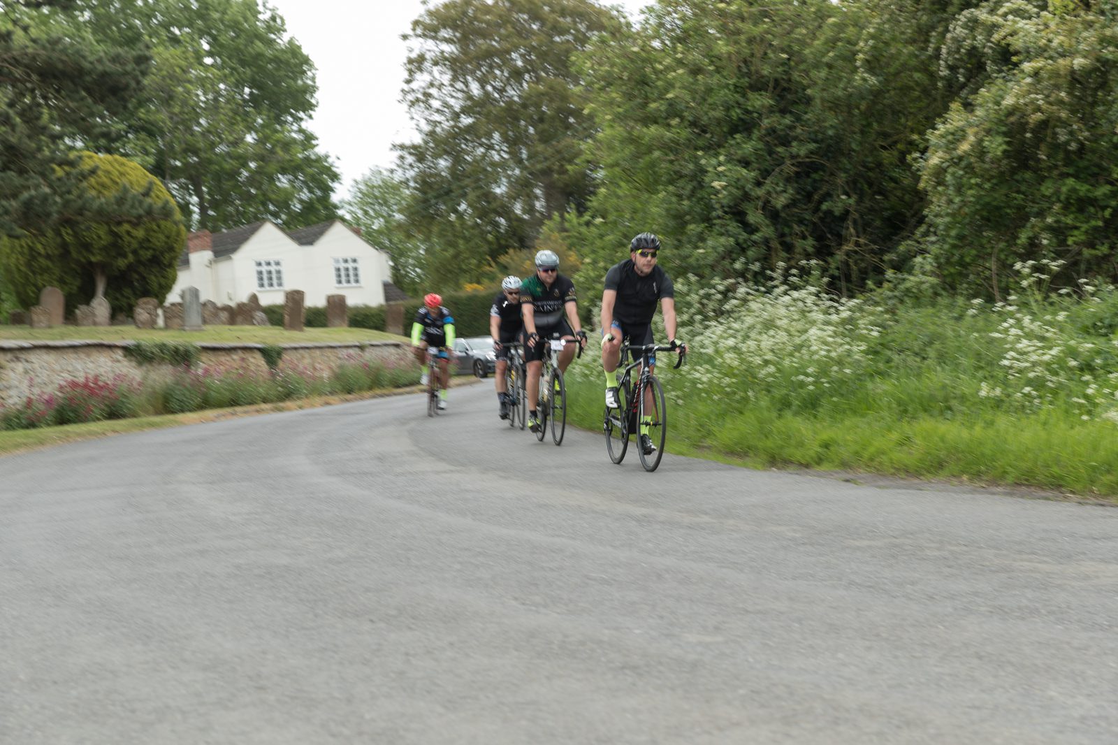 Four cyclists ride on a paved, tree-lined road with greenery on both sides. A white house with a hedge and a stone wall is visible in the background. The cyclists are in a line with the leading rider dressed in black. The scene appears to be calm and leisurely.
