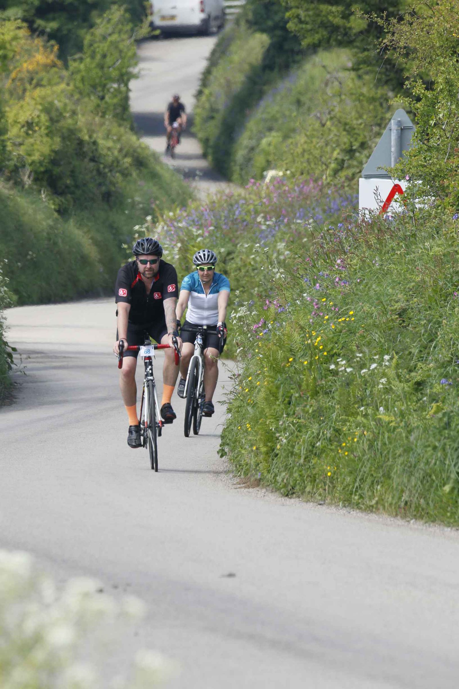 Two cyclists ride on a narrow, uphill country road bordered by lush green foliage and wildflowers. Both wear helmets and cycling gear. The rider on the left is in a black outfit, and the rider on the right is in blue and white. More cyclists can be seen in the distance.