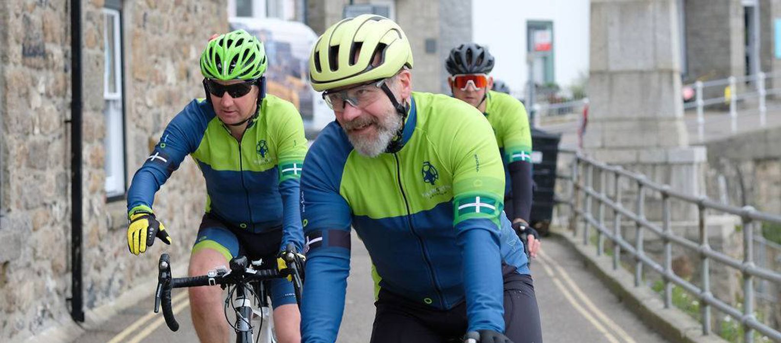 Three cyclists in matching green and blue gear ride along a road near a stone building. They wear helmets and sunglasses, and appear to be enjoying the ride. The setting is urban, with a railing lining the right side of the road.