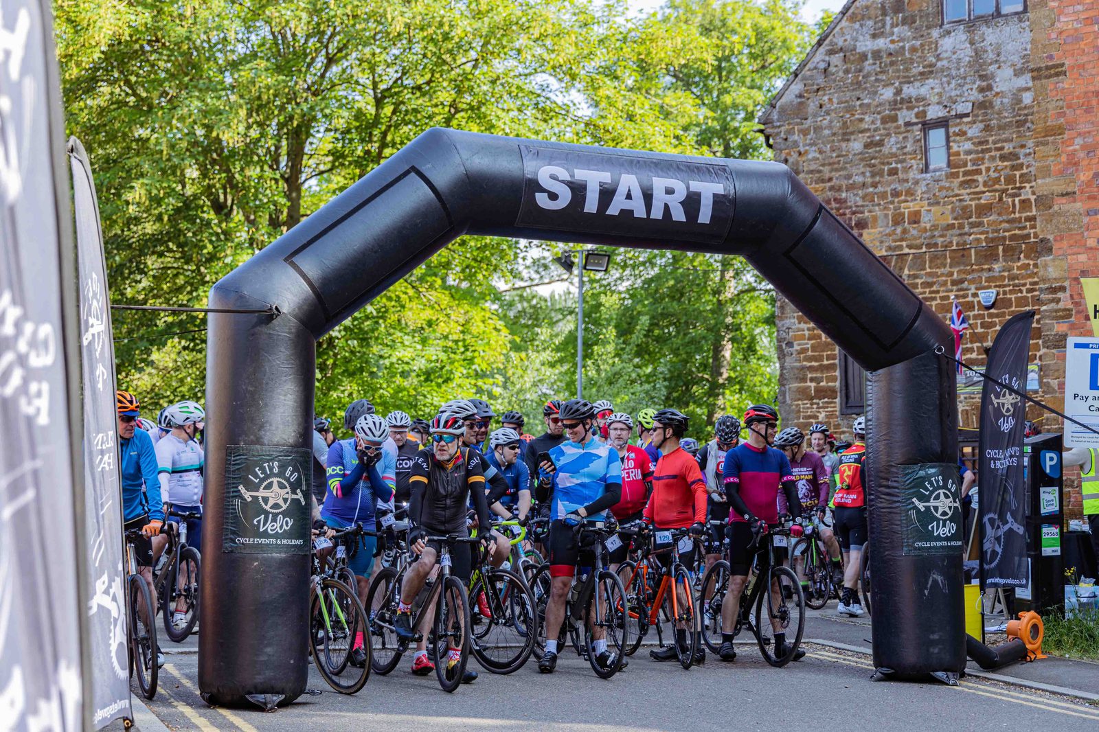 A group of cyclists gathers at the starting line beneath a large black inflatable archway labeled "START." They are ready with their bikes on a sunny day, surrounded by lush green trees and a brick building. Various cycling event banners are visible around the area.