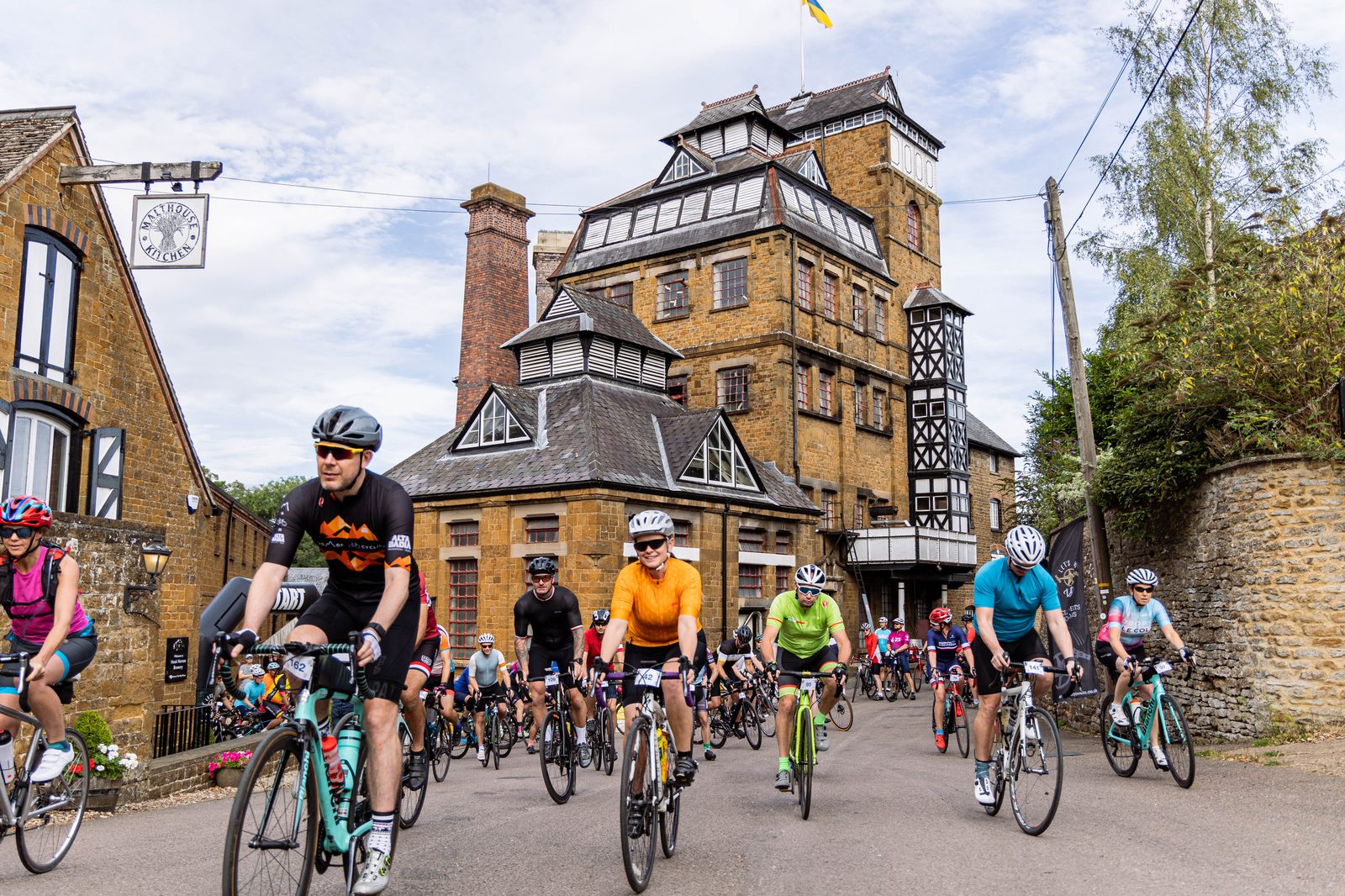 A group of cyclists in colorful attire ride through a village with historic stone buildings and a large brewery in the background. The cyclists wear helmets and sunglasses, and the sky is partly cloudy. The scene depicts a community or race cycling event.