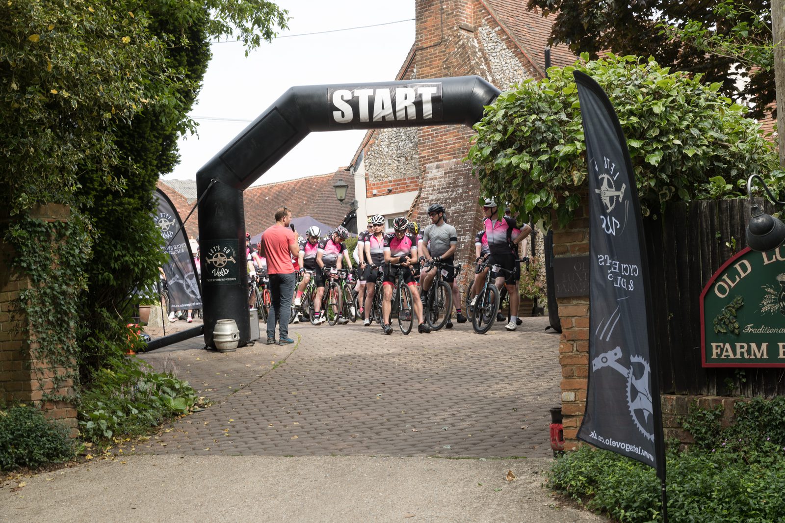 A group of cyclists in matching gear gather at the starting line under a large black balloon arch labeled "START." They prepare to begin a race or event on a paved path surrounded by greenery and rustic buildings. A person in a red shirt stands near the group.