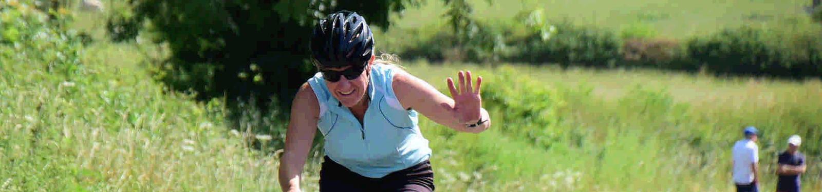 A person wearing a helmet and sunglasses rides a bicycle down a rural path, smiling and waving. Trees and grassy fields line the path, with two other people walking in the distance.