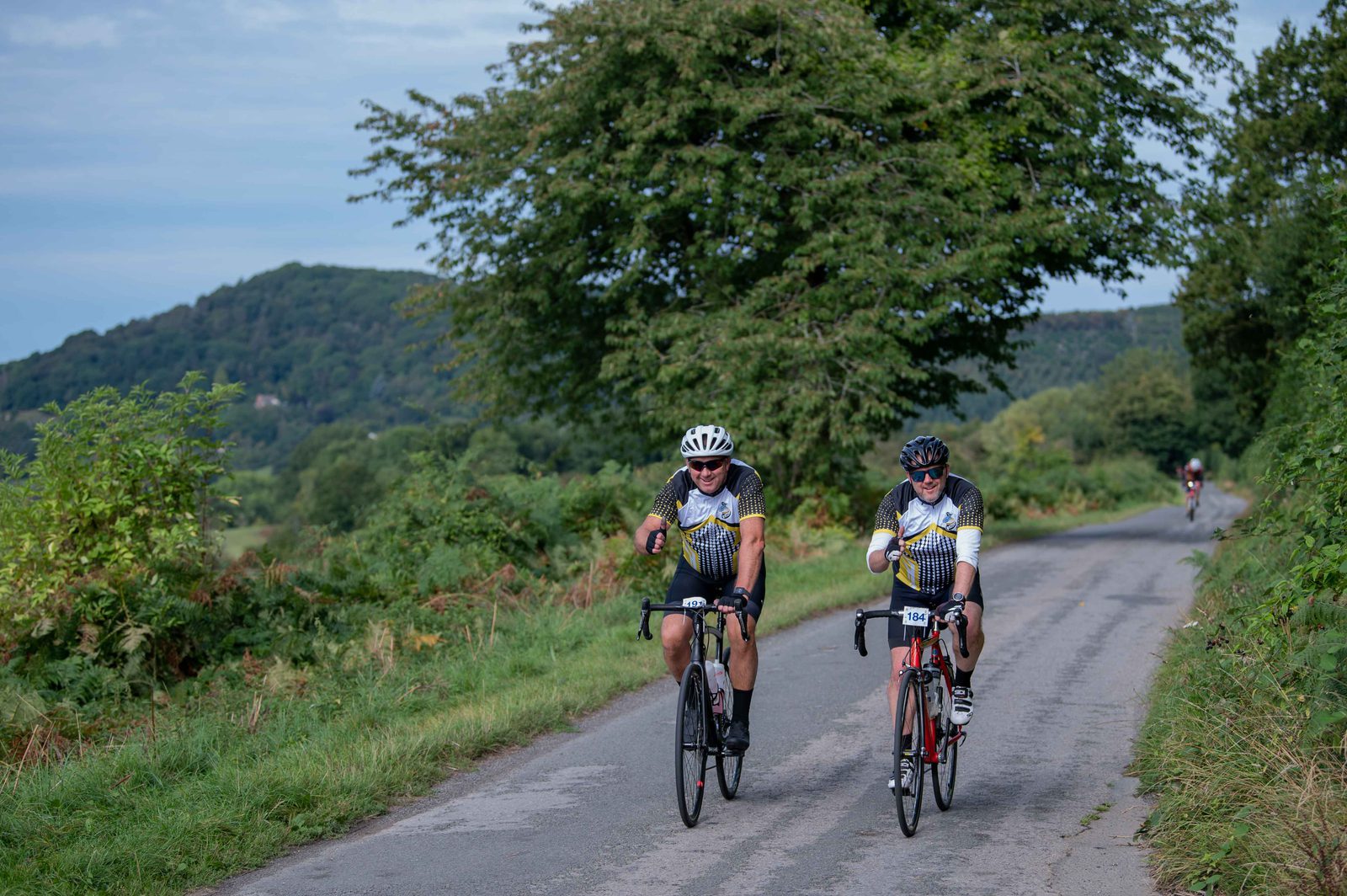 Two cyclists in white helmets and matching jerseys ride on a narrow country road surrounded by greenery and trees. They gesture with thumbs up and are smiling. Hills and a clear sky are in the background, creating a scenic route.