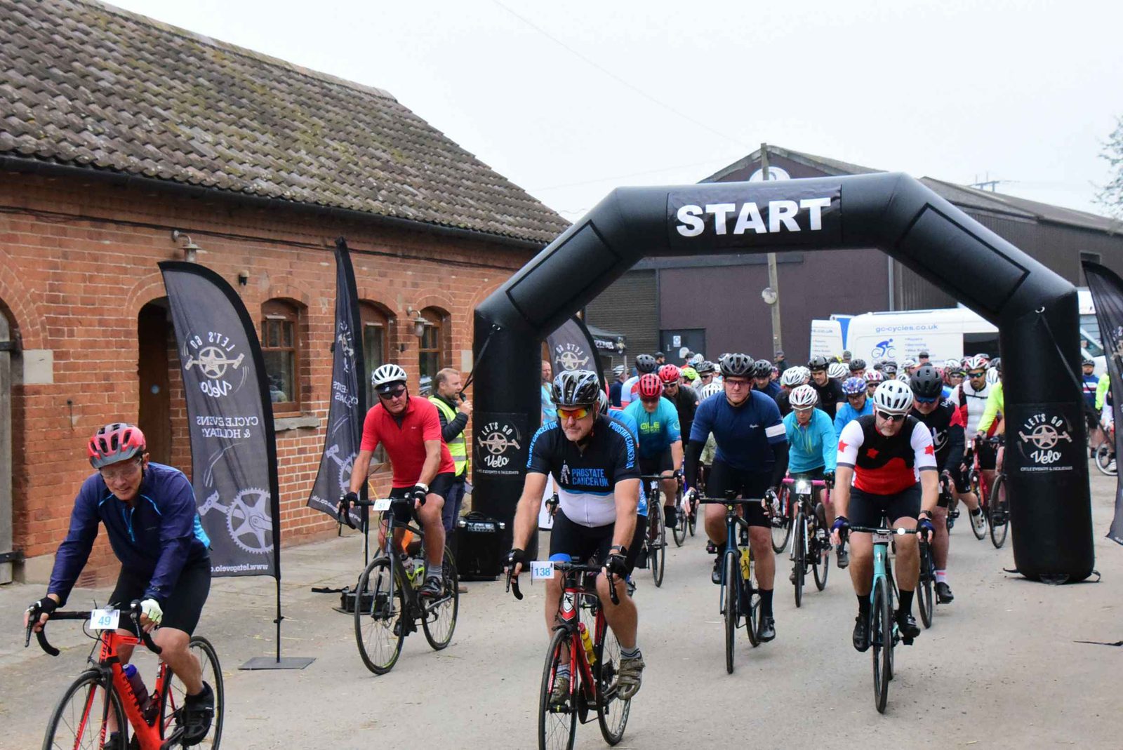 Cyclists in colorful outfits begin a race, riding under a large black inflatable arch with the word "START" on it. They are surrounded by brick buildings and banners displaying race logos. The atmosphere appears energetic and competitive.