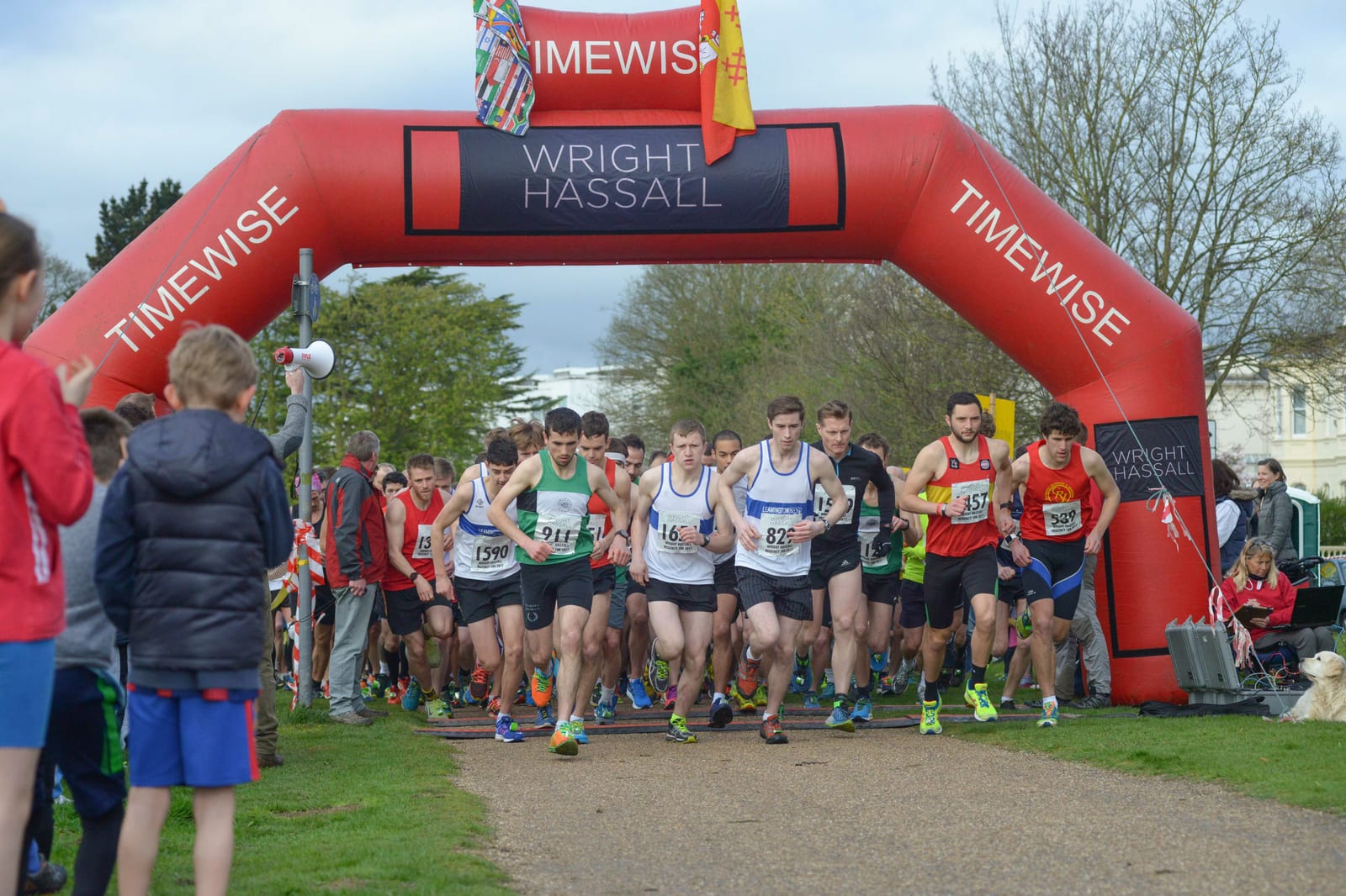 A group of runners in athletic gear start a race at the beginning of a marked path, under an inflatable red archway that reads "TIMEWISE" and "WRIGHT HASSALL." Spectators stand on either side, watching as the runners take off on a grassy, tree-lined course.