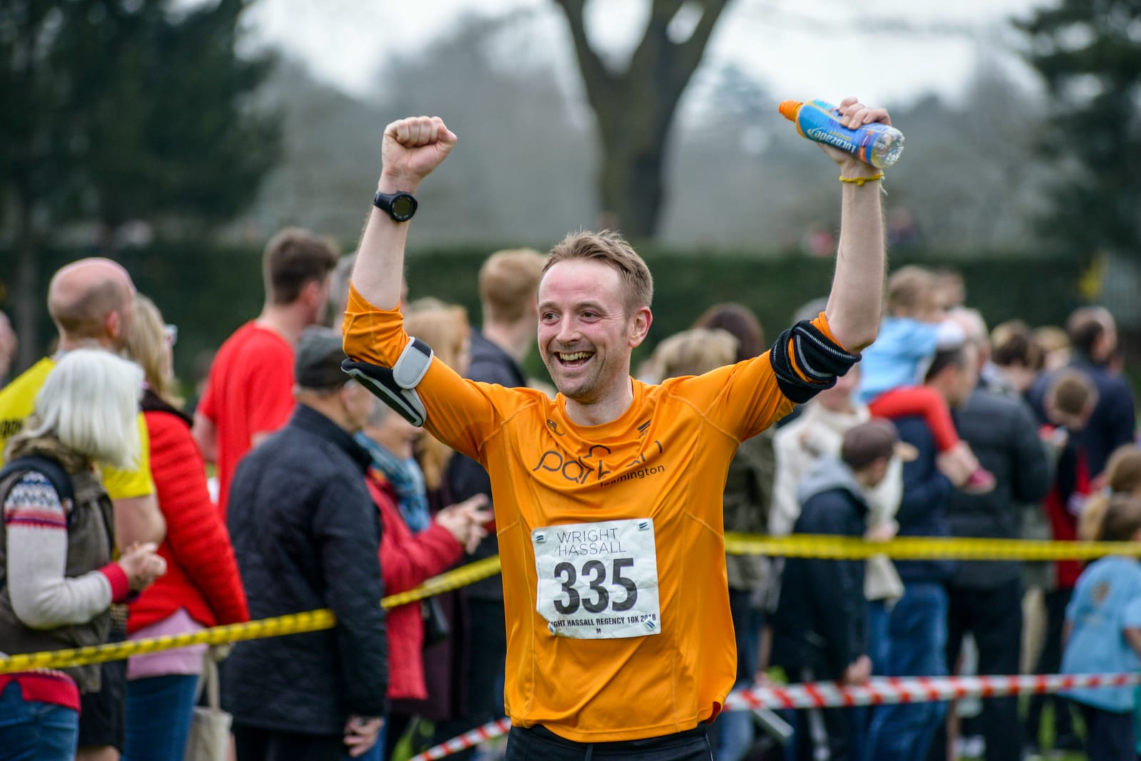 A man in an orange shirt with the number 335 on his chest raises his arms in triumph after finishing a race. He is holding a water bottle in his left hand. Behind him, a crowd of people watches near a yellow barrier tape, with some individuals blurred in the background.