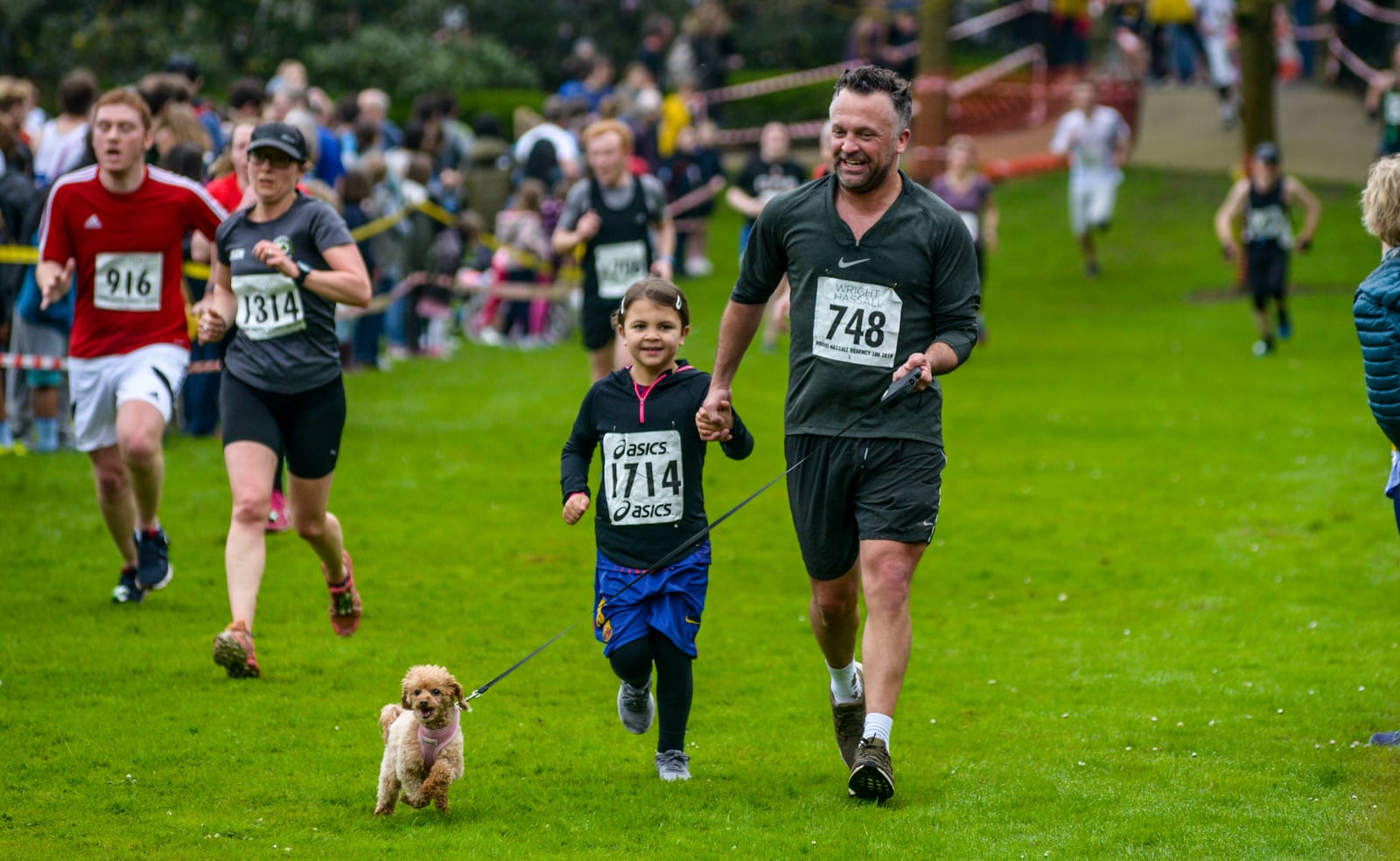 A group of people participating in a race on a grassy field. A man wearing a black outfit with bib number 748 runs alongside a young girl in a black shirt with bib number 174, holding a leash attached to a small dog. Other runners are visible in the background.