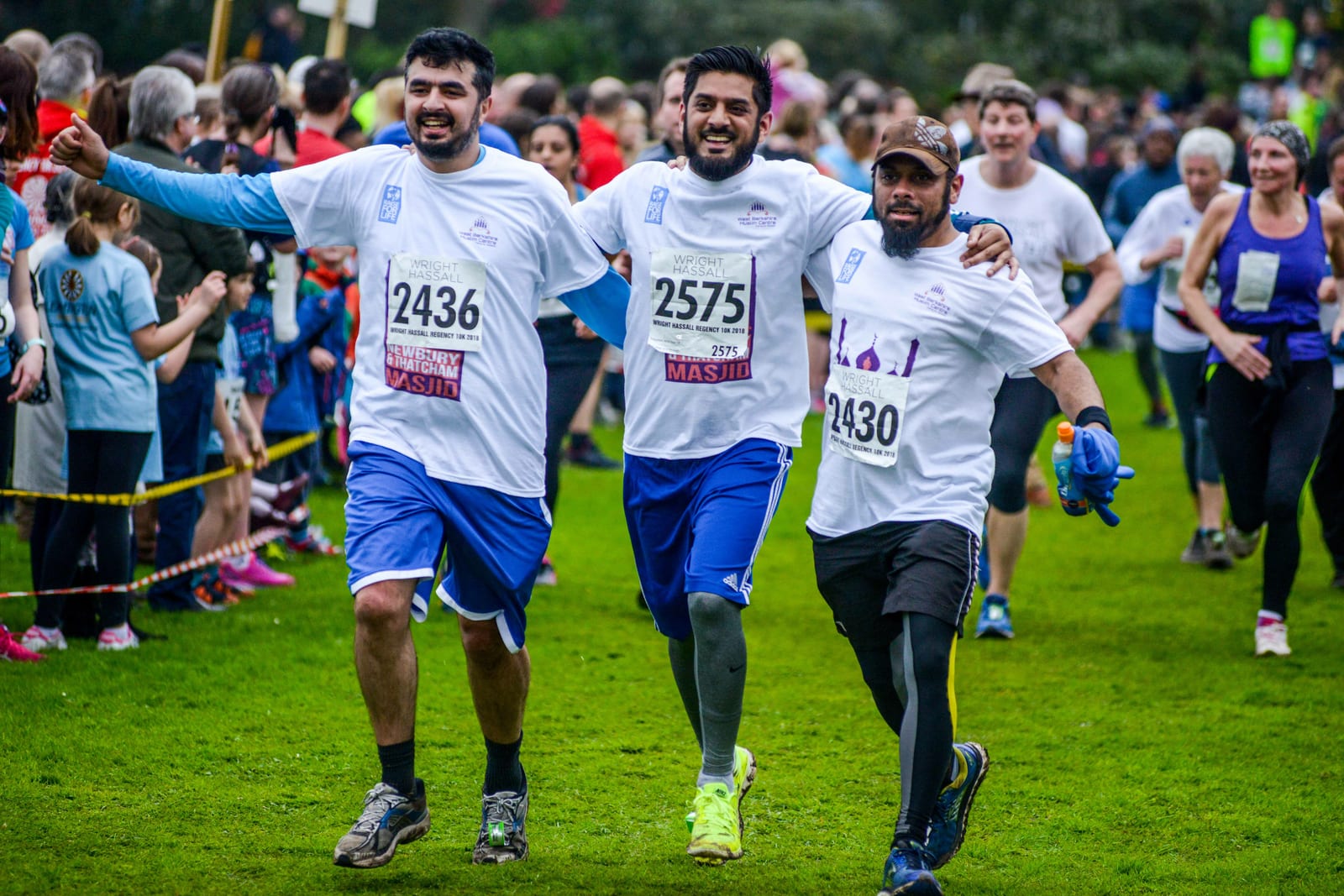 Three men in white T-shirts and shorts, each wearing a race bib, joyfully cross a race finish line together, arms around each other’s shoulders. A crowd of participants and spectators, some in running gear, are visible in the background on a grassy field.