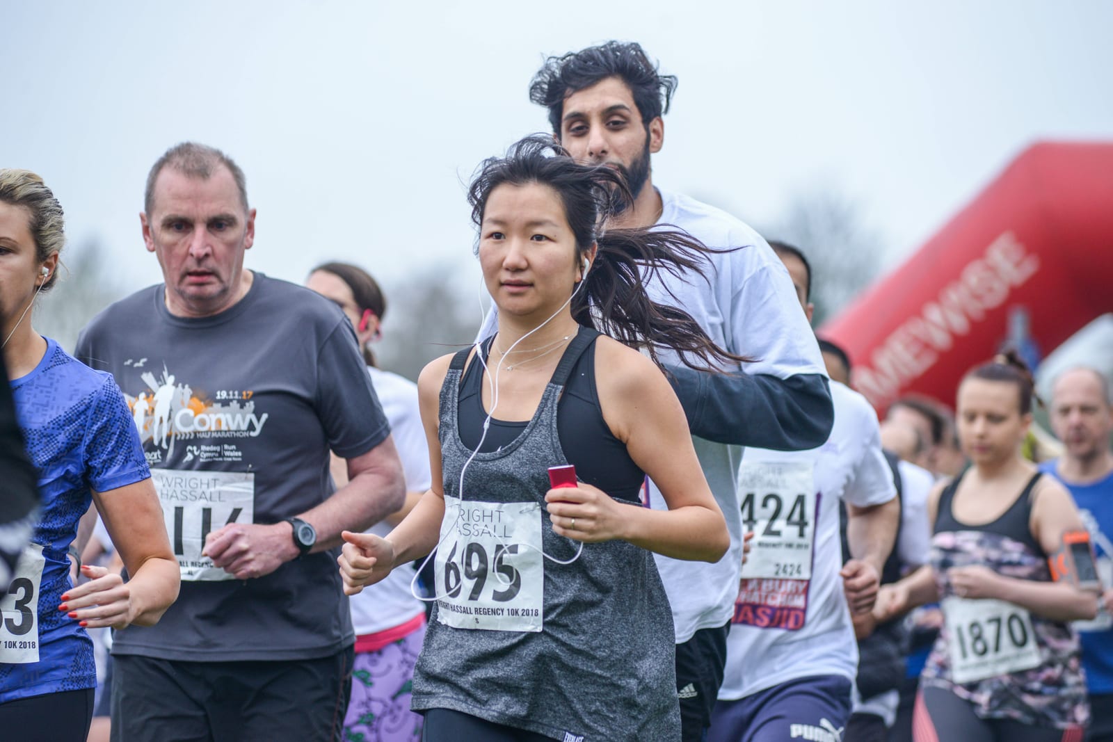 A group of runners participating in a marathon. A woman in the foreground is listening to music through earphones. Runners wear different bib numbers and outfits. The background includes more runners and part of an inflatable arch marking the race pathway.