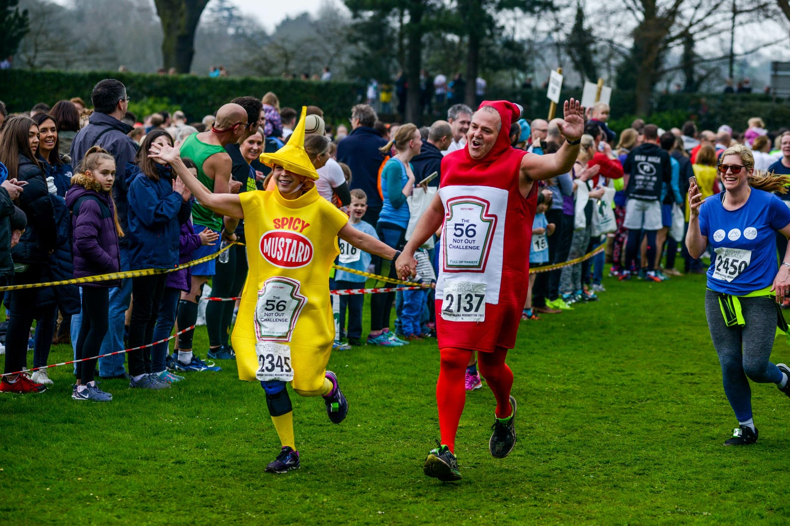 Two people dressed in mustard and ketchup costumes run hand-in-hand during a race, smiling and waving at the cheering crowd. Other runners and spectators are visible in the background. Participants wear numbered bibs and the atmosphere is festive.