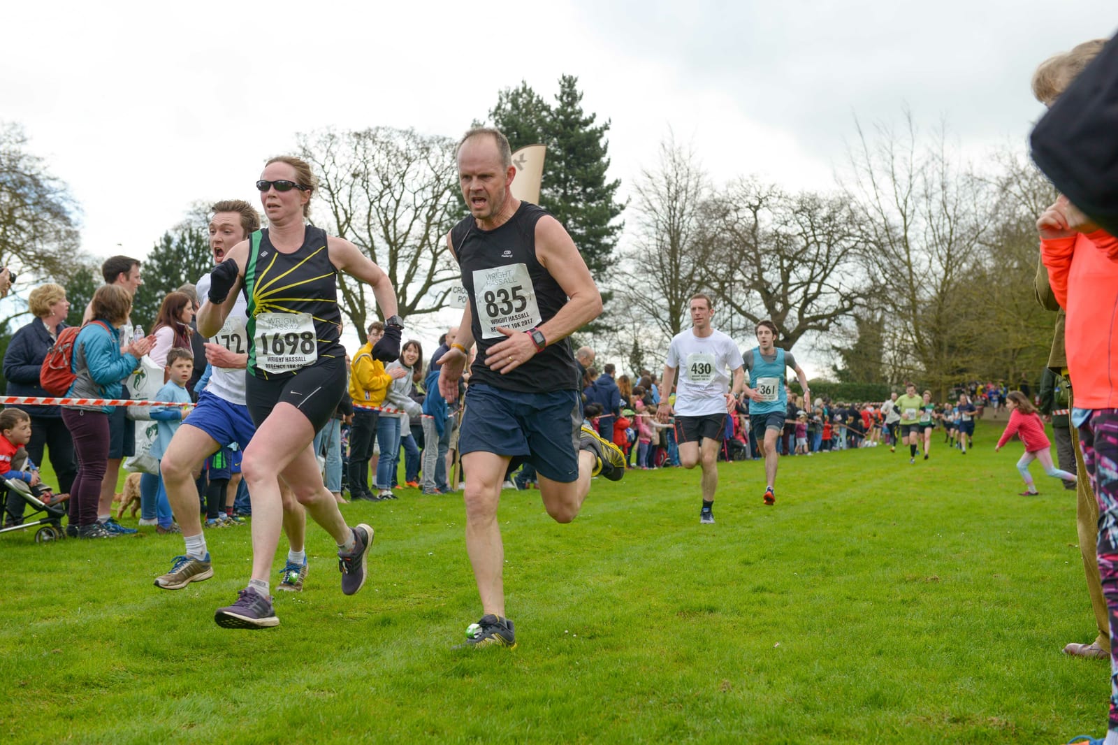 Runners in a race are sprinting on a grassy path with onlookers cheering from the sides. The competitors, wearing numbered bibs, are surrounded by trees and a cloudy sky. Spectators on both sides of the path create a lively atmosphere.