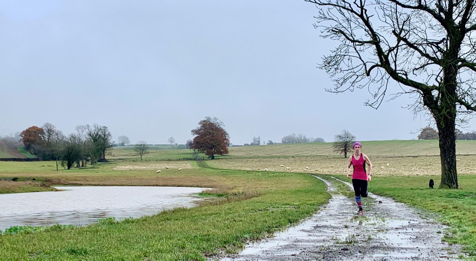 A woman wearing a pink tank top, black leggings, and a pink headband runs along a wet, winding path next to a small lake on a cloudy day. The backdrop features leafless trees, open fields, and a peaceful, overcast sky.