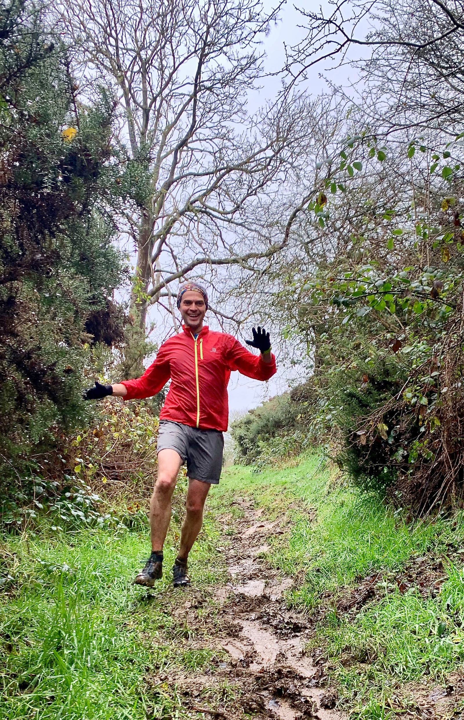 A person wearing a red jacket and gray shorts is running on a muddy trail in a forested area. They are smiling, with both hands raised mid-air, enjoying the outdoor activity despite the cloudy weather. The path is surrounded by trees and bushes.