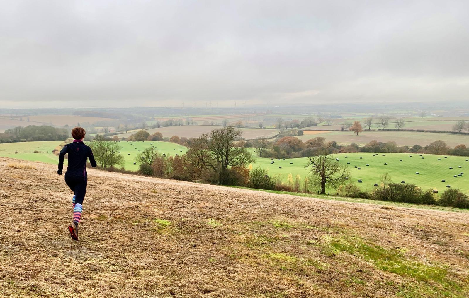 A person wearing a black outfit and colorful striped socks runs across a grassy hill. The background shows a picturesque landscape with rolling green fields, scattered trees, and hay bales under a cloudy sky.