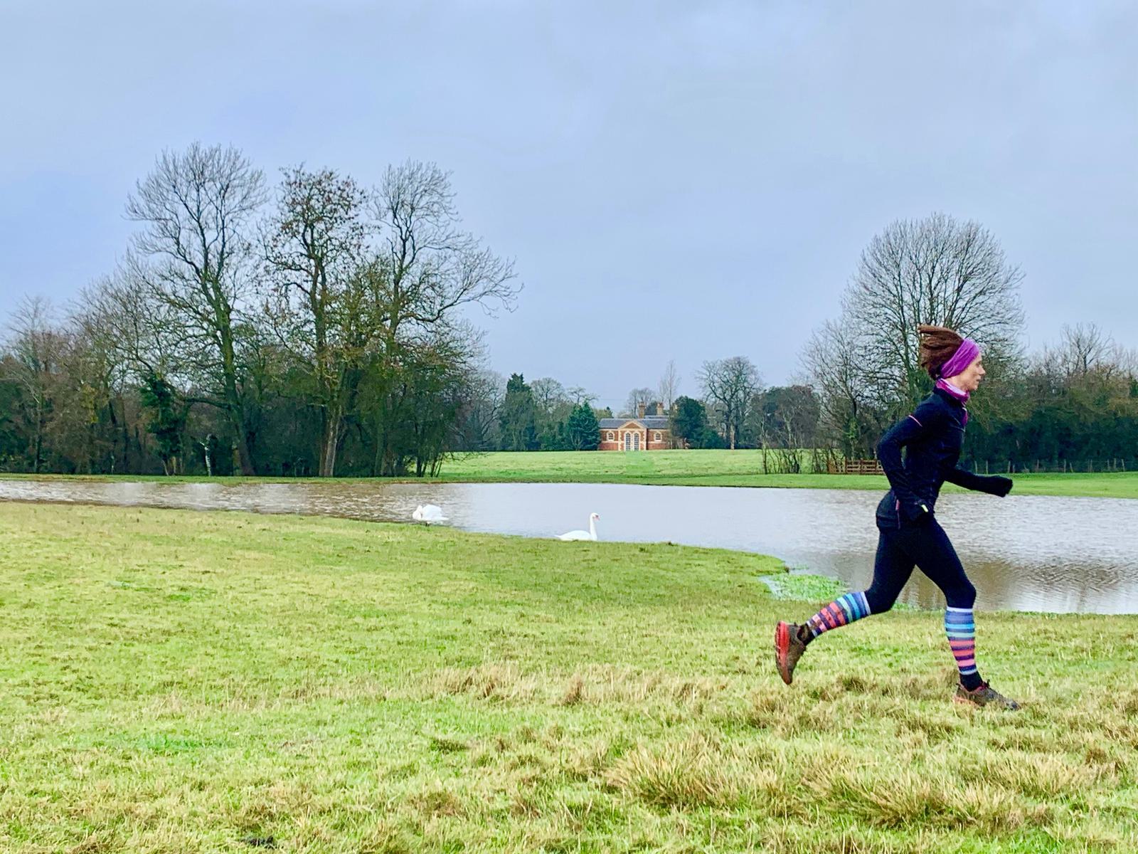 A person is jogging on a grassy field near a body of water, with trees and a building visible in the background. They are dressed in dark athletic wear with colorful striped leg warmers and a hat. A white swan is swimming in the water.