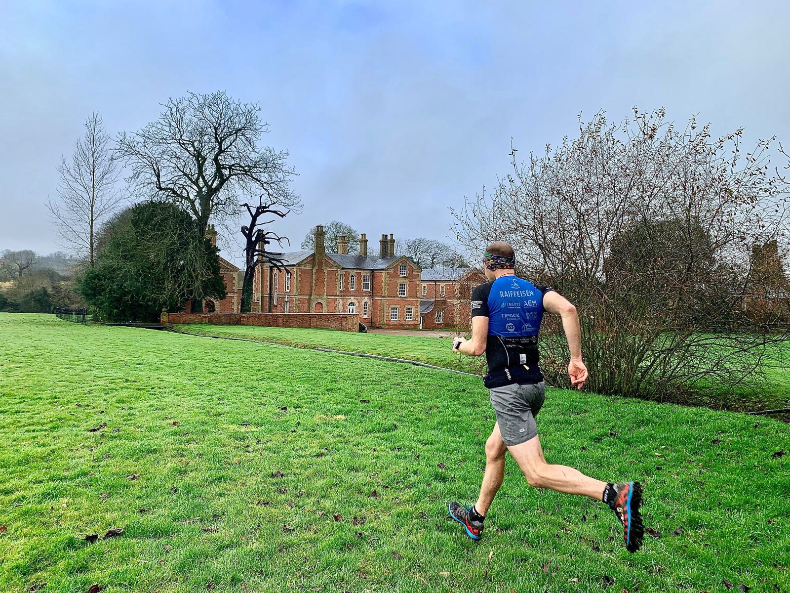 A person wearing a blue shirt and grey shorts is running across a grassy field with a large brick building and leafless trees in the background. The sky is overcast, and the landscape appears to be part of a large estate or park.