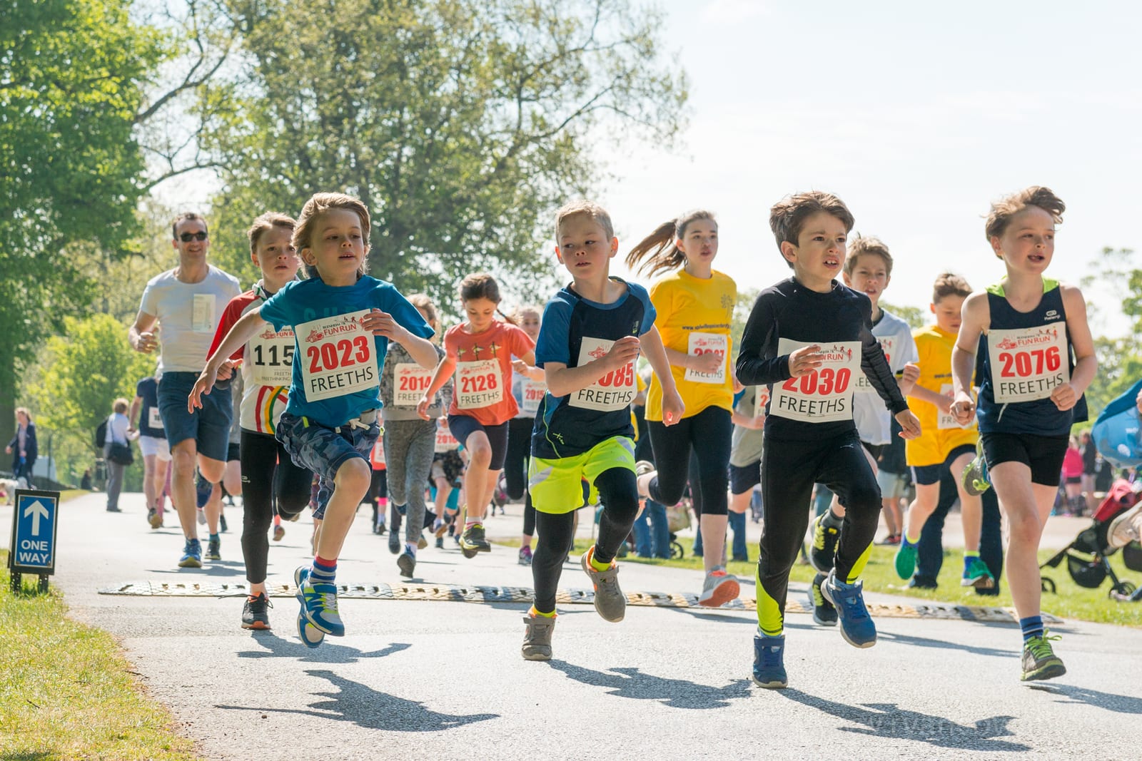 A group of children are running in a race outdoors on a sunny day. They are wearing running gear with numbered bibs pinned to their shirts. Spectators and trees are visible in the background, adding to the lively atmosphere of the event.