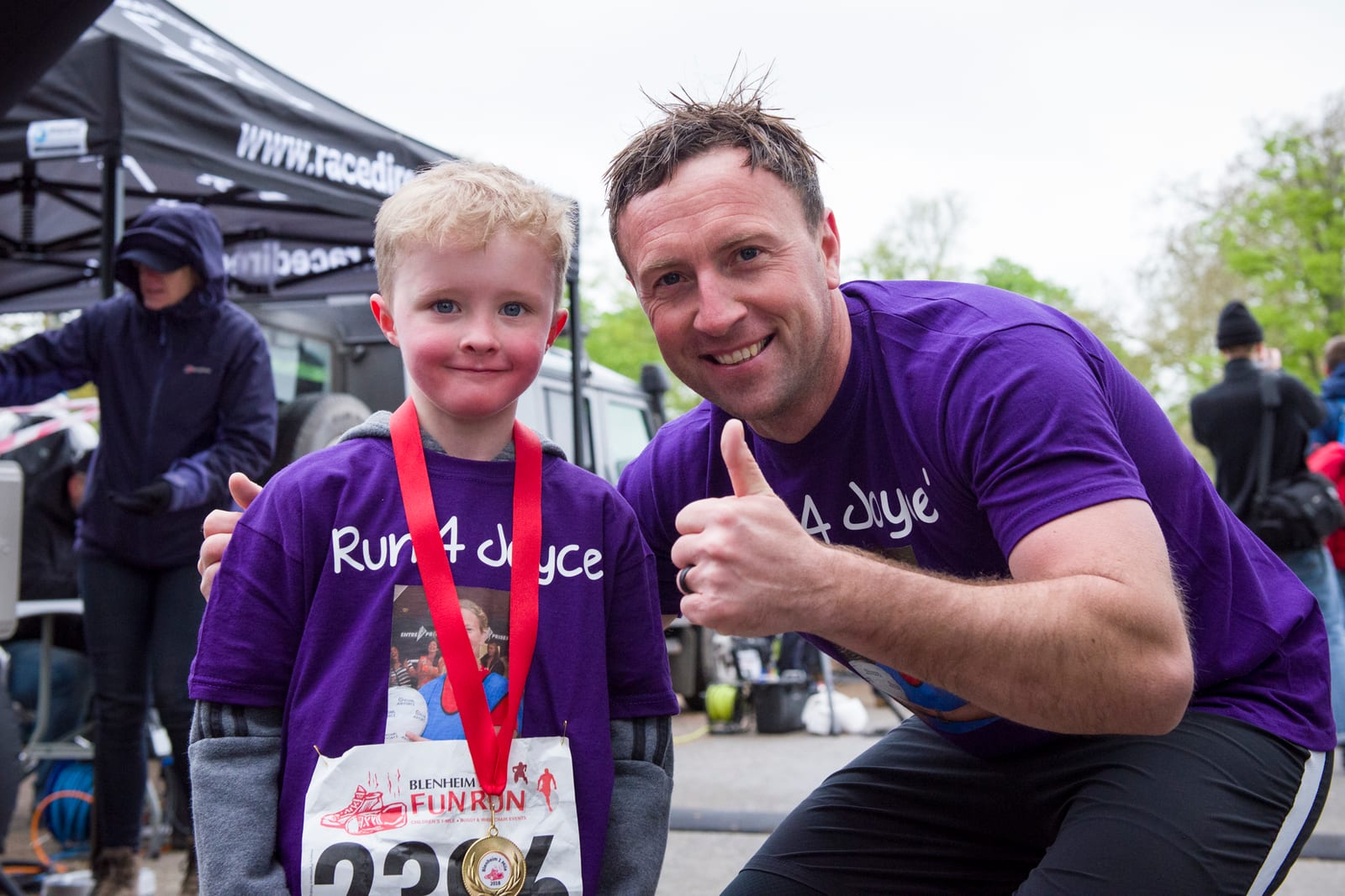 A young boy and an adult, both wearing purple "Run 4 Joye" T-shirts, pose for a photo at a fun run event. The boy, wearing race number 296, has a red medal around his neck, while the adult gives a thumbs-up. Tents and other participants are visible in the background.