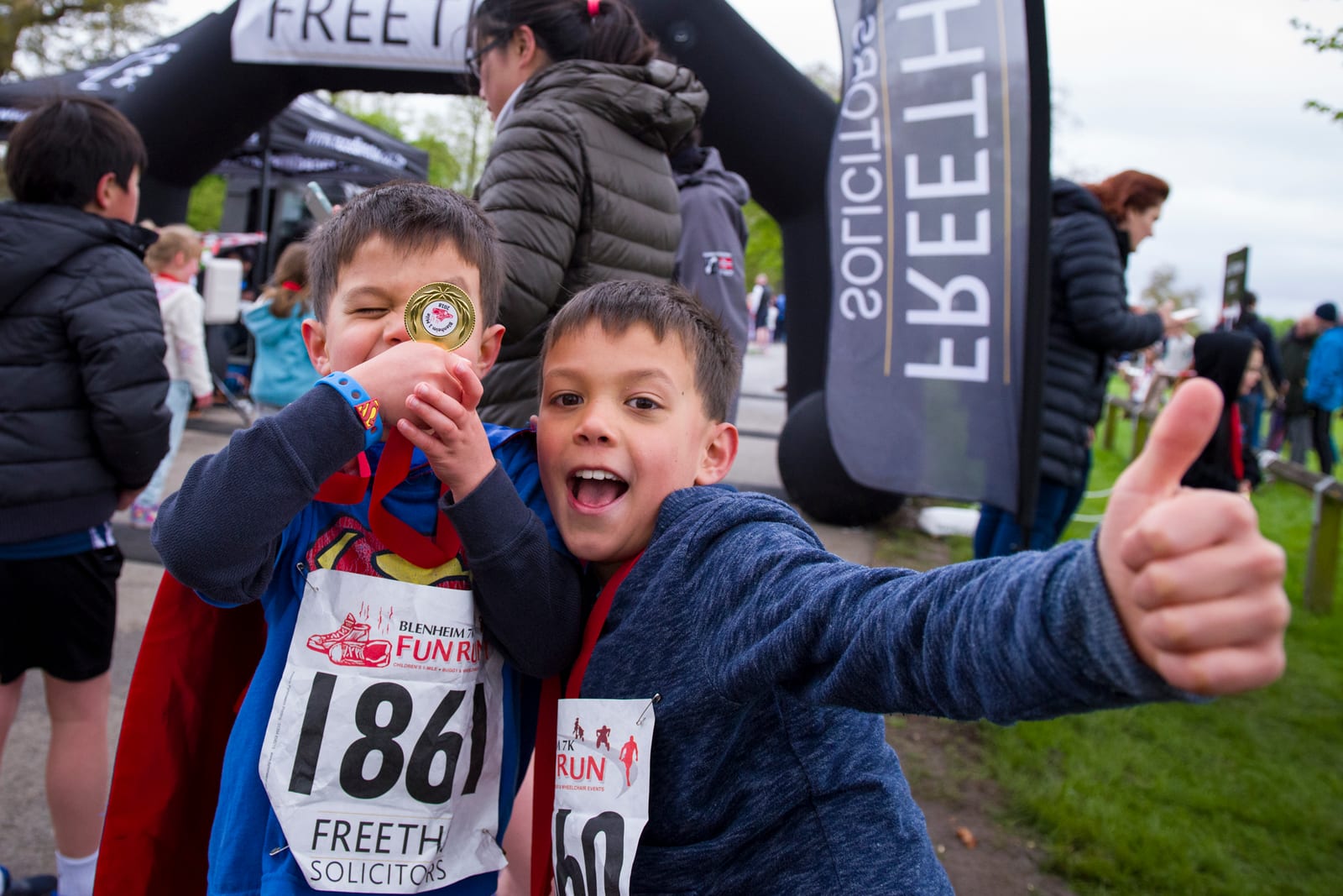 Two young boys pose enthusiastically at a fun run event, with race bibs numbered 186 and 160. The boy on the left proudly displays a medal and wears a Superman shirt, while the boy on the right, in a blue hoodie, gives a thumbs-up. A "FREETH Solicitors" banner is in the background.
