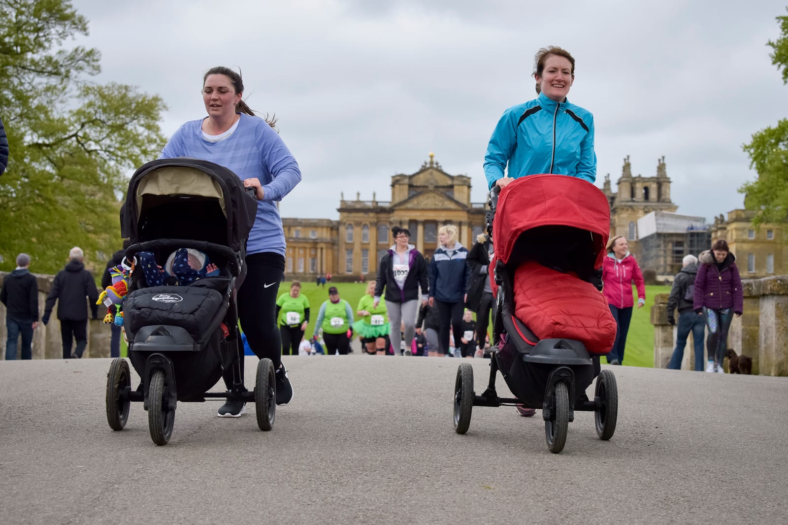 Two women push strollers while participating in a race event on a paved pathway with a historic building in the background. The woman on the left wears a purple top, and the woman on the right wears a blue jacket. Other participants are visible behind them.