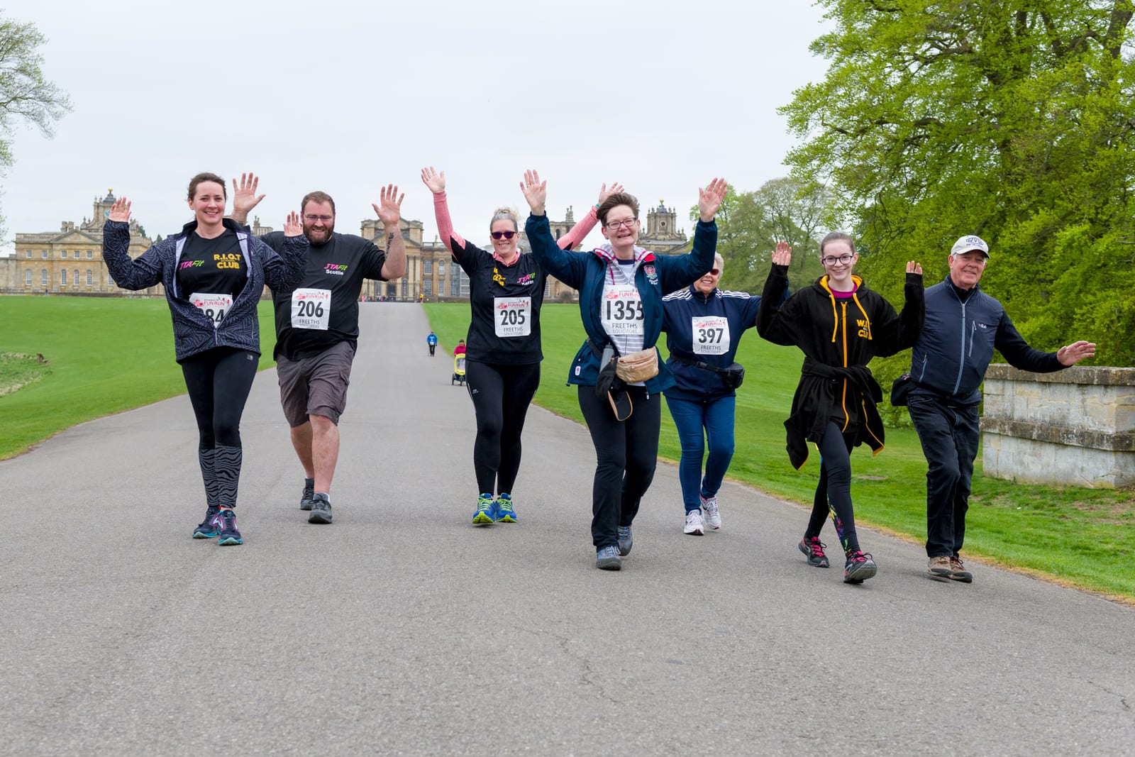 A group of seven people is participating in a running event, with race numbers pinned to their outfits. They are smiling and waving as they walk or jog along a paved road with greenery and buildings in the background.