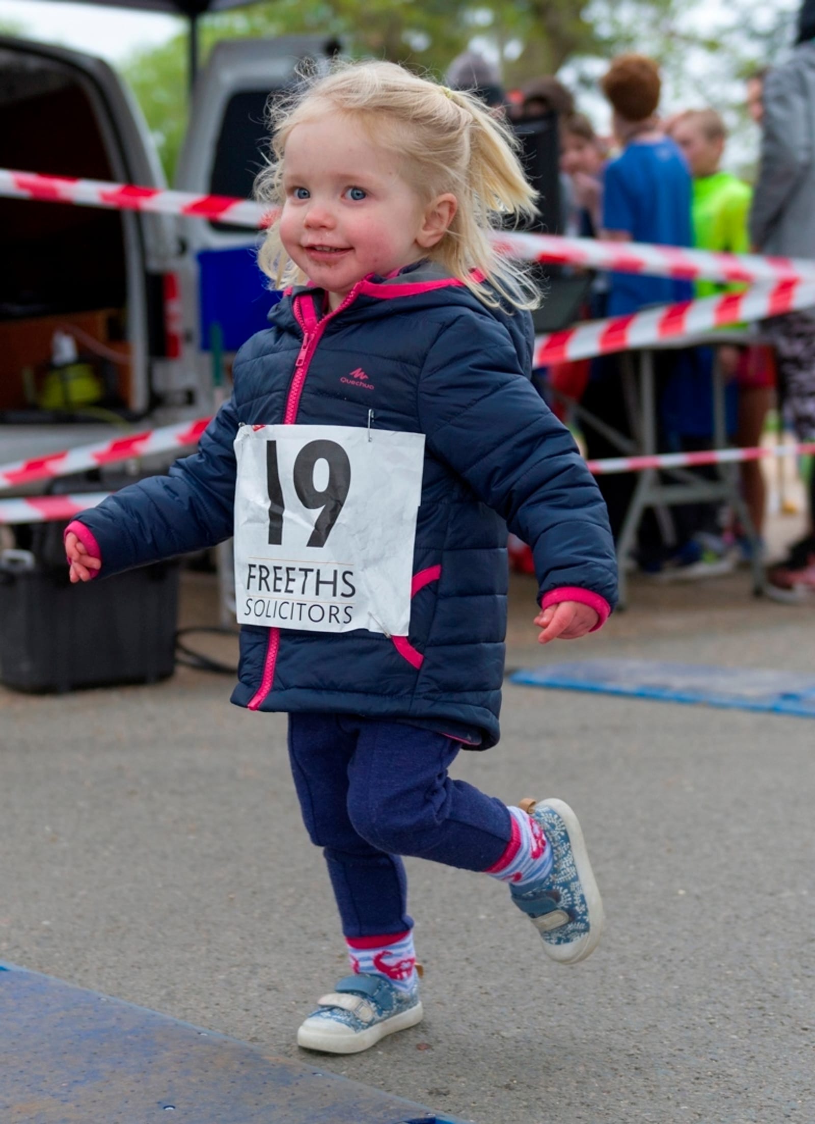 A young girl with blond hair, wearing a black jacket with pink accents, blue pants, and sneakers, participates in a race. She wears a bib number 19 and smiles as she runs on a pavement, with a crowd and red-and-white tape in the background.