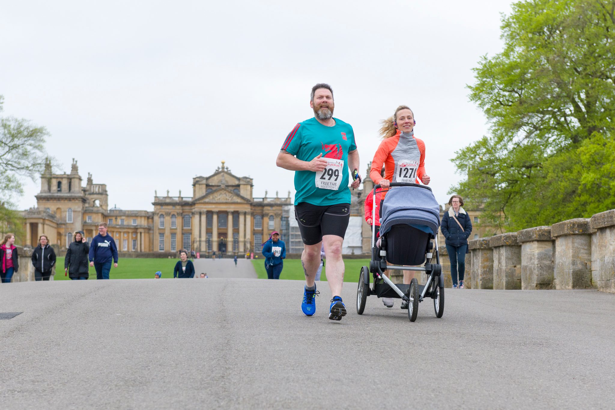 A man and a woman, both wearing race bibs and athletic attire, run together on a paved path with a stroller in front of a large, historic building. Other participants can be seen in the background, along with a few trees and spectators to the sides.