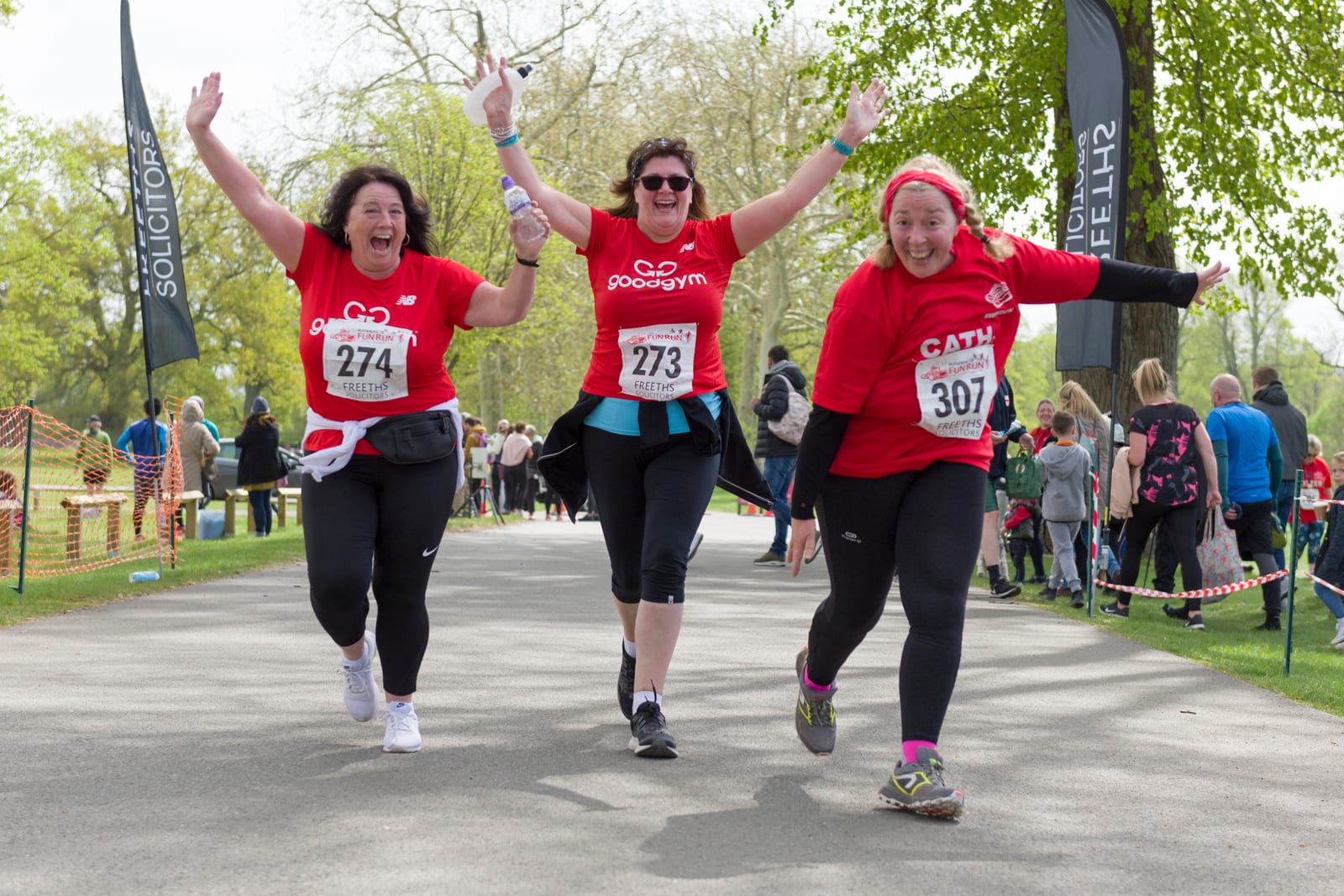 Three women in athletic attire joyfully run hand-in-hand on a park pathway during a race. All are wearing "GoodGym" shirts, with running numbers 274, 273, and 275. Spectators are visible in the background near trees and flags.