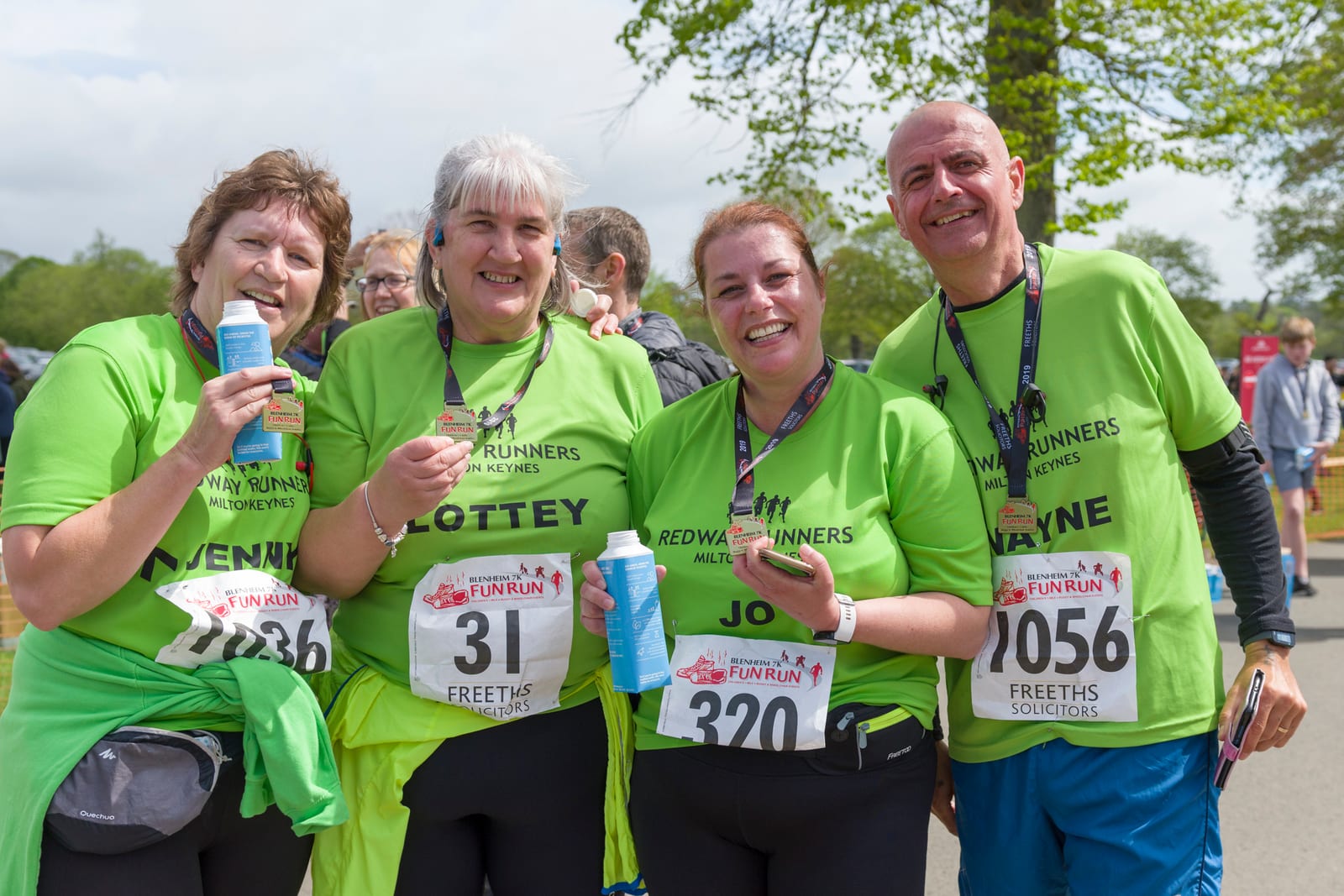 A group of four people, wearing matching neon green "Redway Runners" shirts, pose together for a photograph after a fun run event. They are smiling and holding bottles of water and snacks. They each have race bibs with different numbers pinned to their shirts.