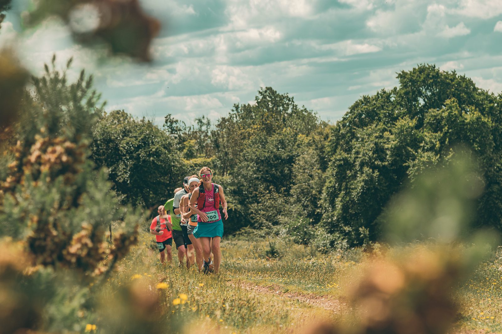 Four people run on a grassy trail surrounded by trees and bushes under a partly cloudy sky, participating in an outdoor race on a sunny day.