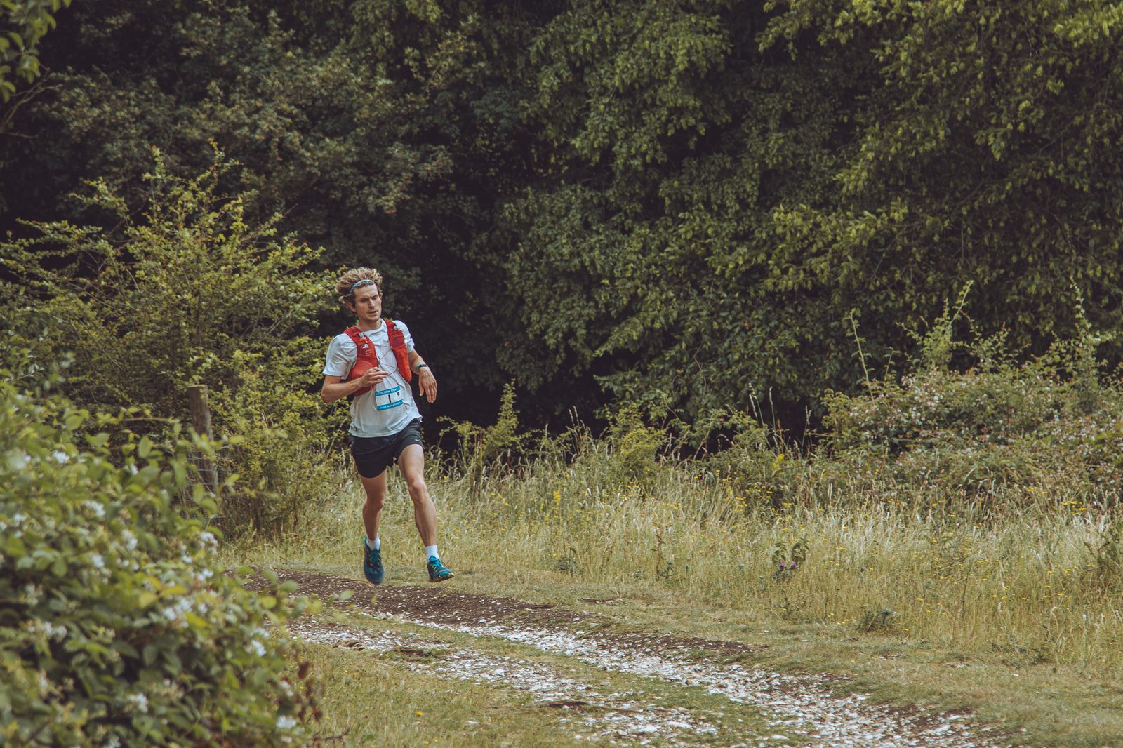 A man in athletic gear and a race bib runs on a dirt trail surrounded by greenery and dense trees, participating in an outdoor race or marathon.