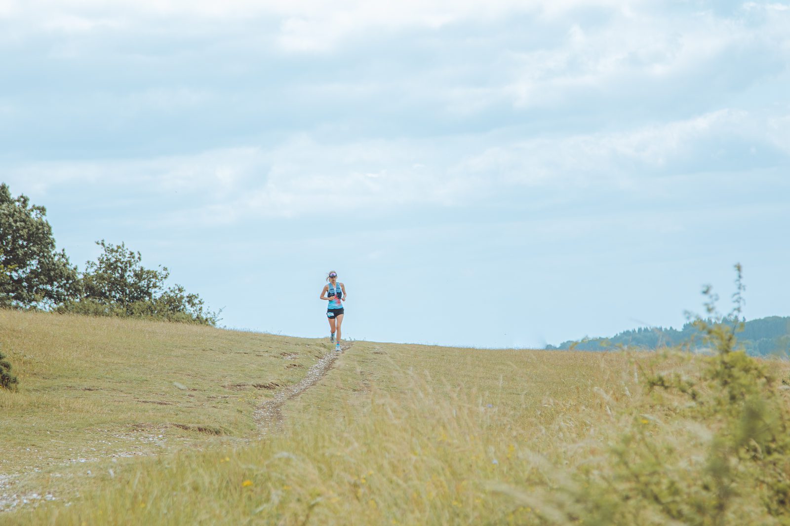 A person in athletic gear is running along a narrow dirt path in an open grassy field. The sky is mostly cloudy, and the landscape appears to be rural with sparse trees and distant greenery.