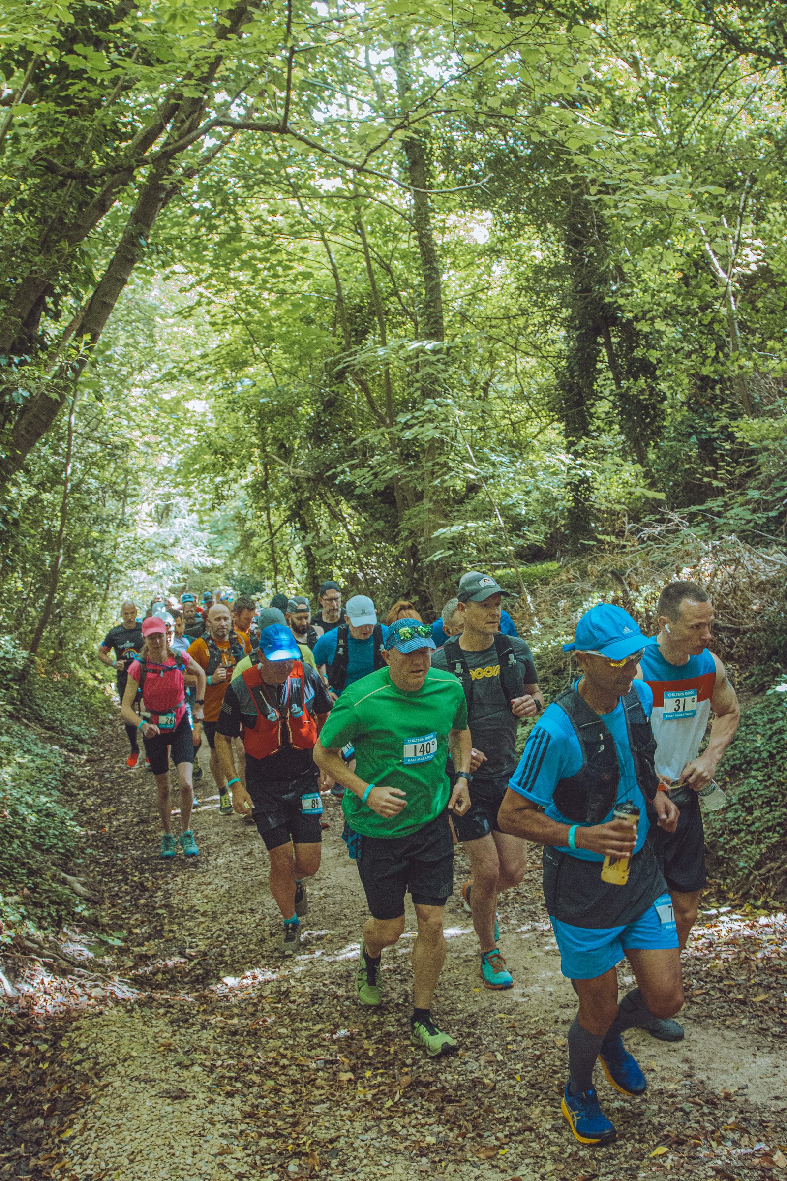 A group of runners wearing colorful athletic gear and race bibs run together through a forest trail. Sunlight filters through the dense canopy of trees, illuminating the path and casting dappled shadows on the ground. The runners appear focused and determined.
