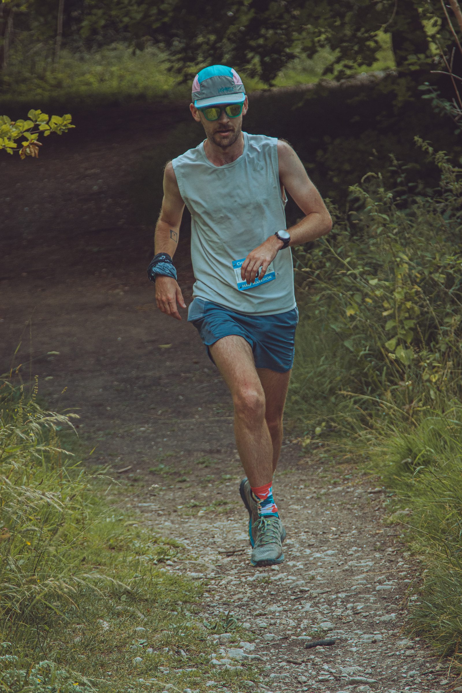 A person wearing a blue hat, sunglasses, a light gray sleeveless shirt, blue shorts, and running shoes jogs on a forest trail. They have a blue wristband and a smartwatch on one arm, and the trail is surrounded by green foliage.