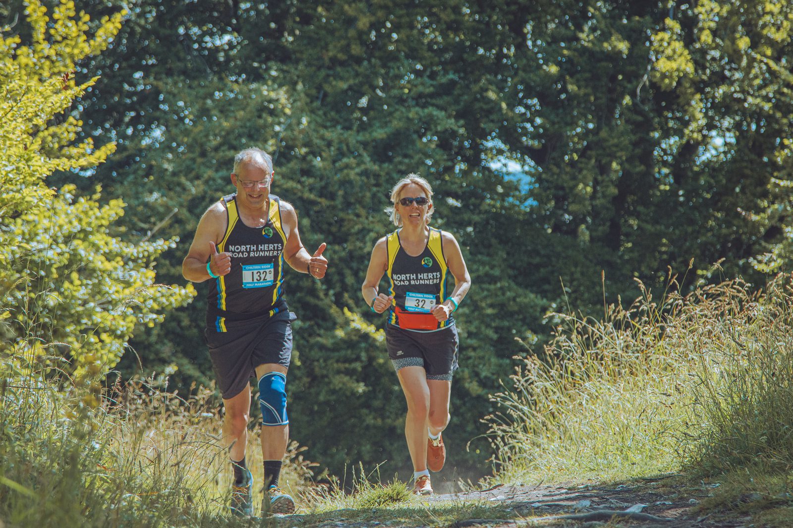 Two runners, wearing "North Herts Road Runners" vests, run along a sunlit, tree-lined path. The male runner, with a blue knee brace, gives a thumbs up, while the female runner smiles. Both have race numbers pinned to their shirts and appear to be mid-race.
