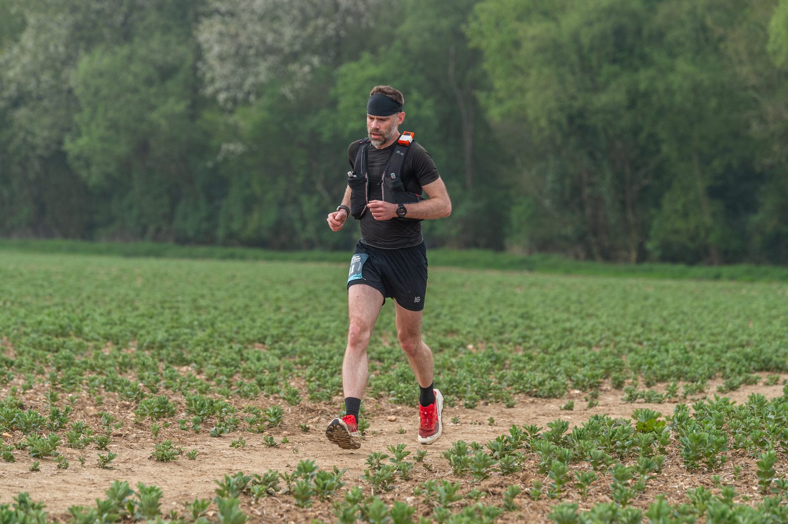 A man in athletic gear runs on a dirt path through a green field, surrounded by vegetation and trees. He wears a headband, backpack, race bib, and bright red running shoes.