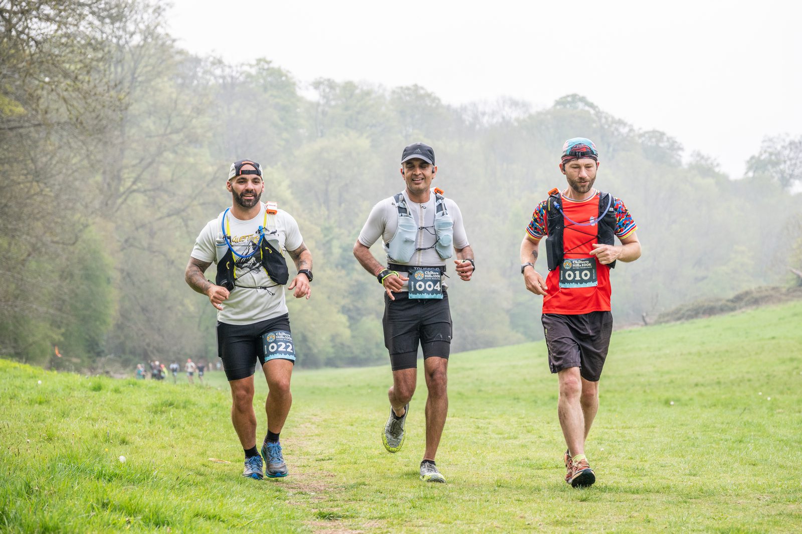 Three men are running on a grassy trail outdoors, wearing race bibs and athletic gear. Trees and more runners are visible in the background on a foggy day.