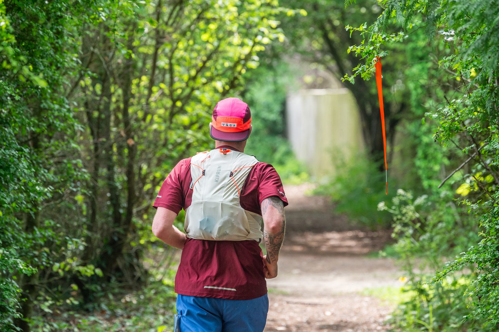 A person wearing a red cap, red shirt, blue shorts, and a white hydration pack runs along a sunlit dirt trail surrounded by green trees and bushes, with an orange marker hanging from a branch.