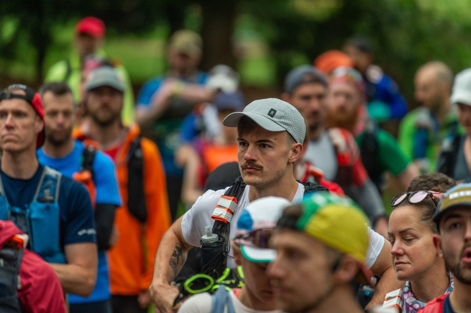 A group of runners, mostly men, stand closely together outdoors, wearing athletic gear and hydration packs. The focus is on a man with a mustache in a light gray cap, looking ahead with concentration.