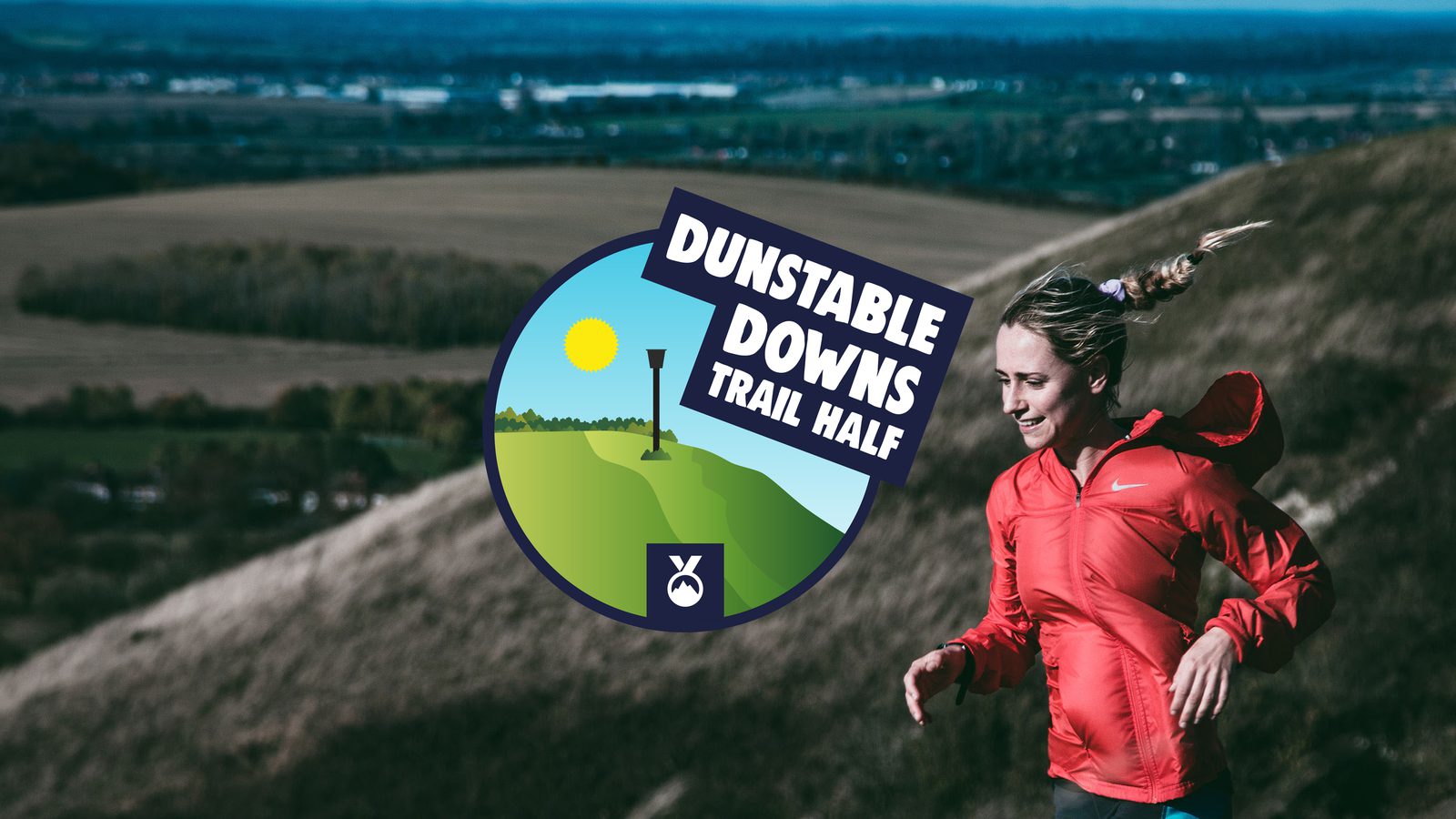 A woman in a red jacket runs on a trail with scenic hills in the background. Overlay text reads "Dunstable Downs Trail Half," featuring a logo with a sun, hill, trail marker, and medal icon. The background showcases sprawling fields and a distant horizon.