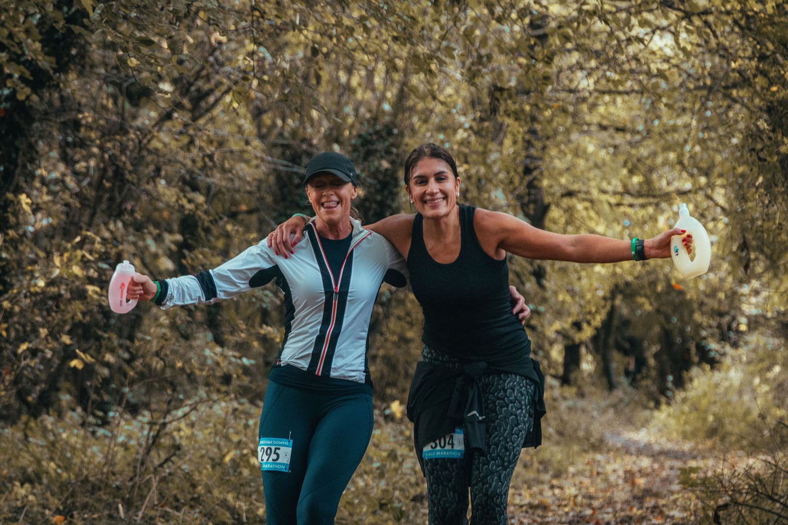 Two women joyfully running through a forest trail, wearing athletic gear and holding drink containers. They have their arms around each other's shoulders and are smiling. Race numbers are visible on their outfits.