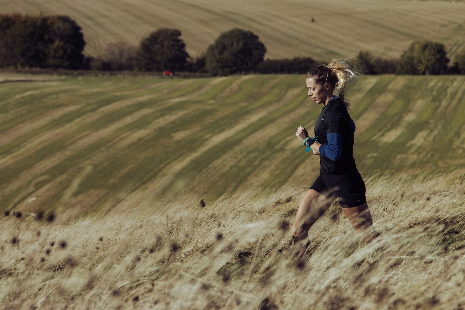 A person with long hair tied back is running uphill through a grassy field. They are wearing athletic clothing and appear focused on their activity. The background features expansive, rolling farmland with some trees scattered in the distance.