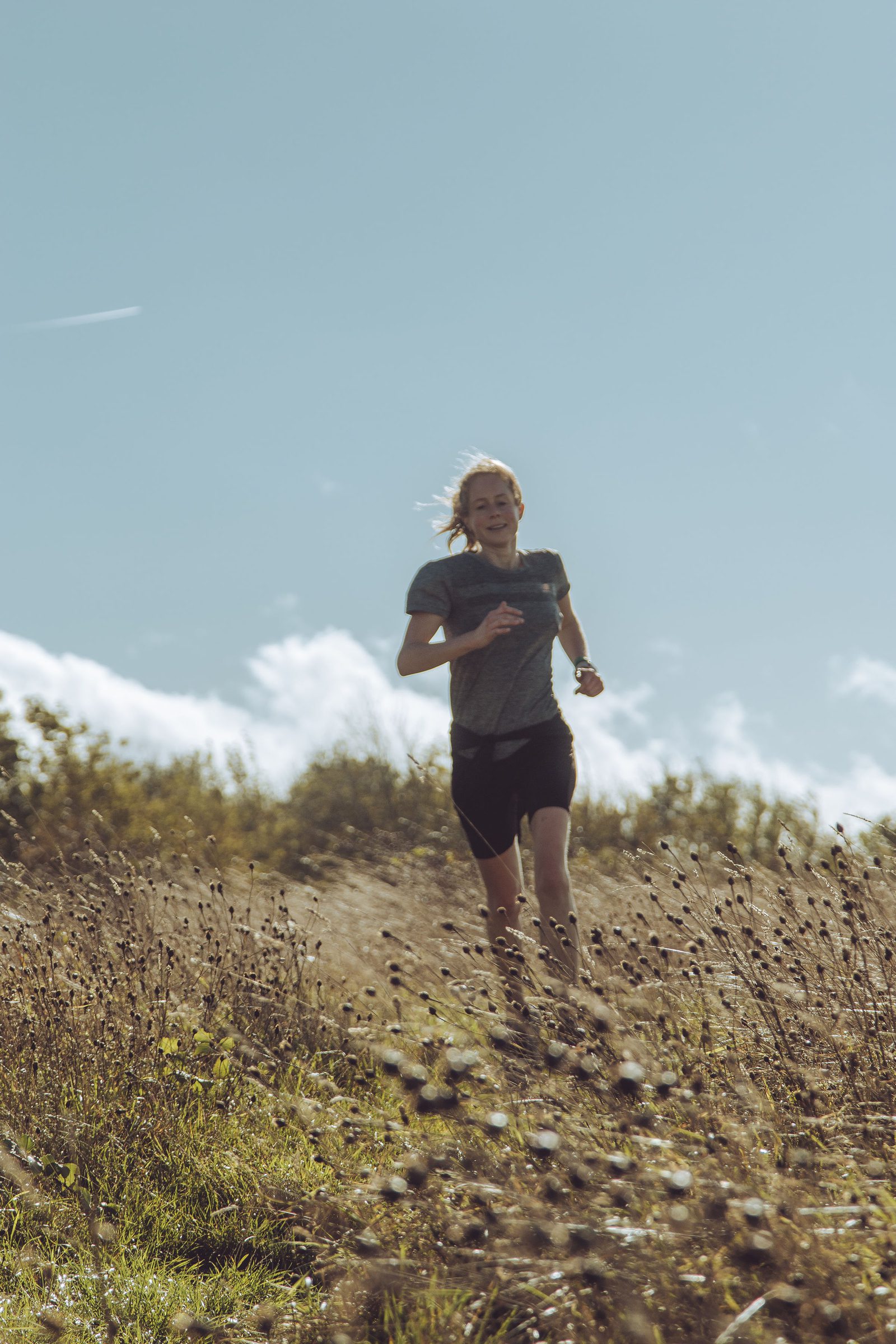 A person with long hair is running outdoors through a grassy field on a sunny day. The sky is clear and blue, and the person is wearing a grey shirt and black shorts. Bushes and trees can be seen in the background.
