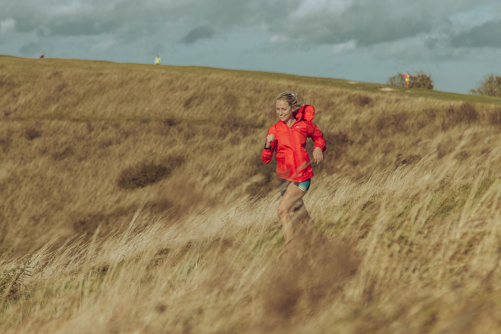 A person with long hair runs through a grassy field on a windy day, wearing a bright orange jacket and shorts. The sky is overcast with clouds, and the field stretches into the distance. Other individuals are visible in the background at the top of the hill.