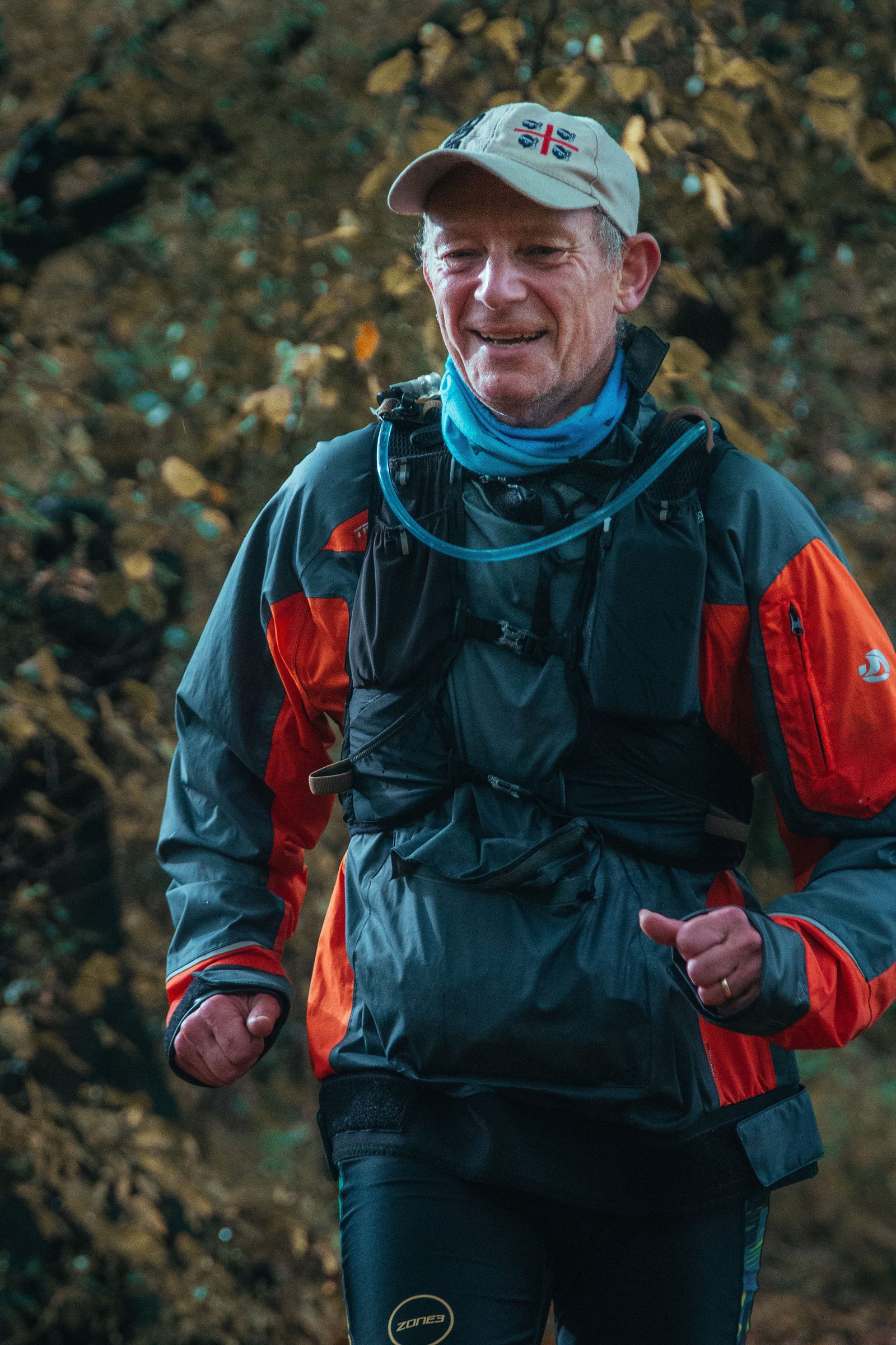 A man wearing a beige cap, a neck gaiter, and a red-and-black jacket is jogging outdoors. He has a black hydration pack on his chest and is smiling. The background is blurred with green foliage.