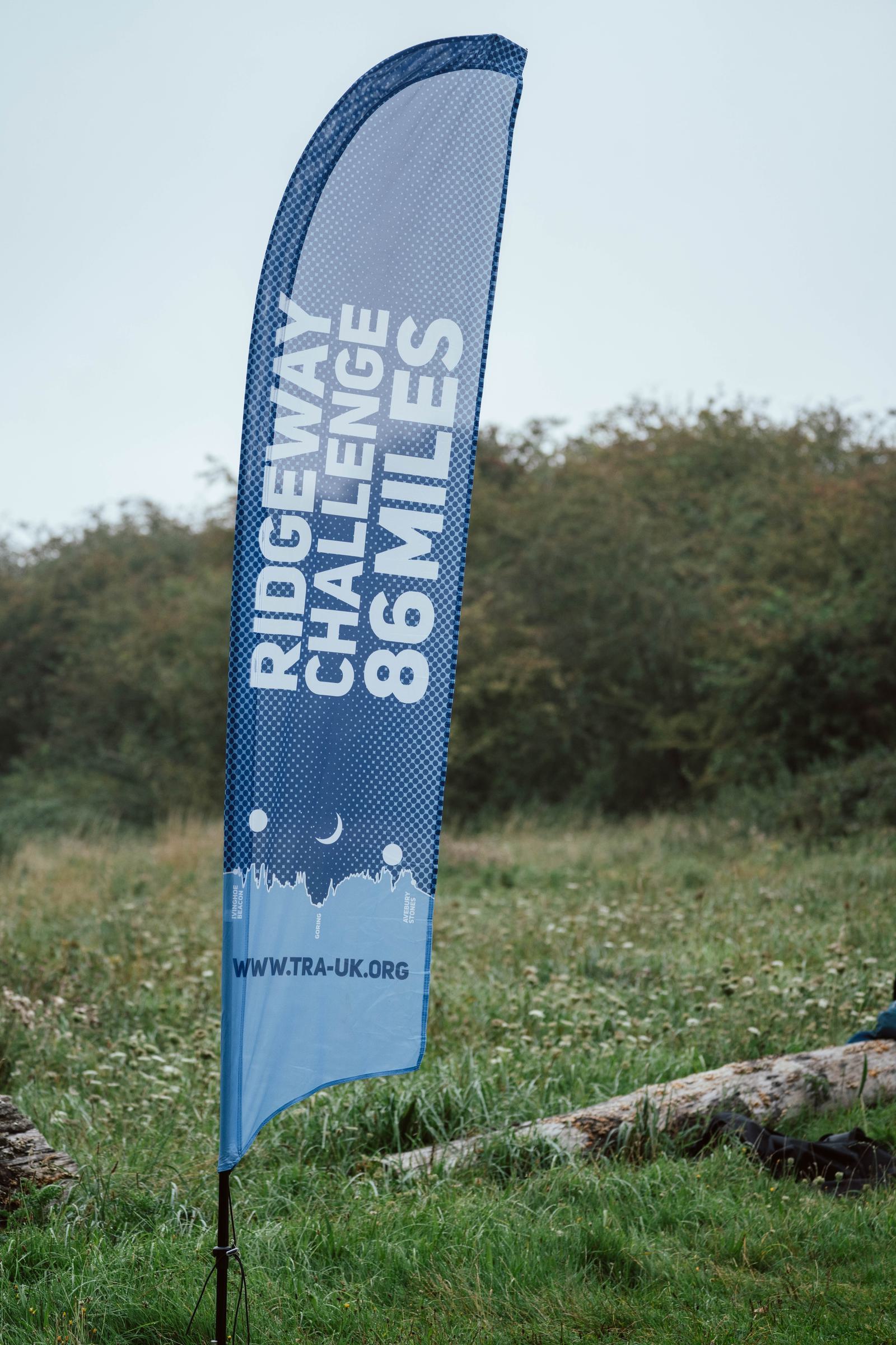 A vertical banner stands outdoors on a grassy field, displaying text that reads "Ridgeway Challenge 86 Miles." The bottom part of the banner shows a silhouette of trees and includes the website "www.tra-uk.org." Trees and bushes are visible in the background.