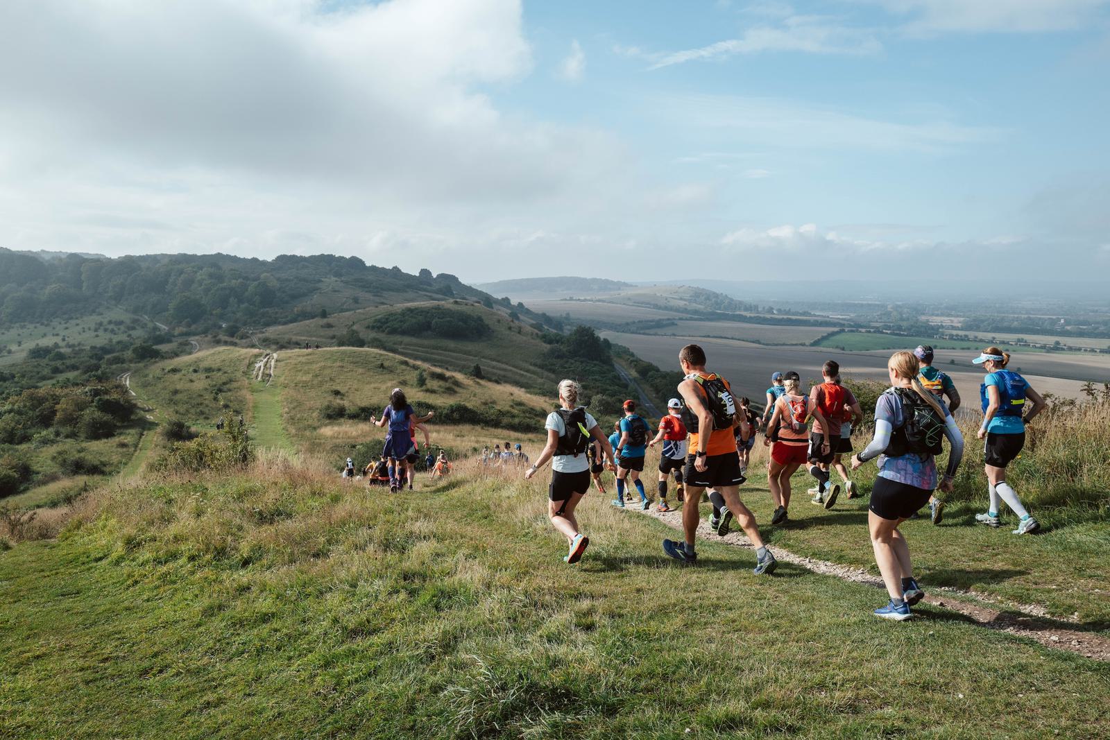 A group of people are running along a rugged trail on a grassy hill with vast countryside scenery in the background. The sky is partially cloudy, and the runners are dressed in vibrant athletic gear.