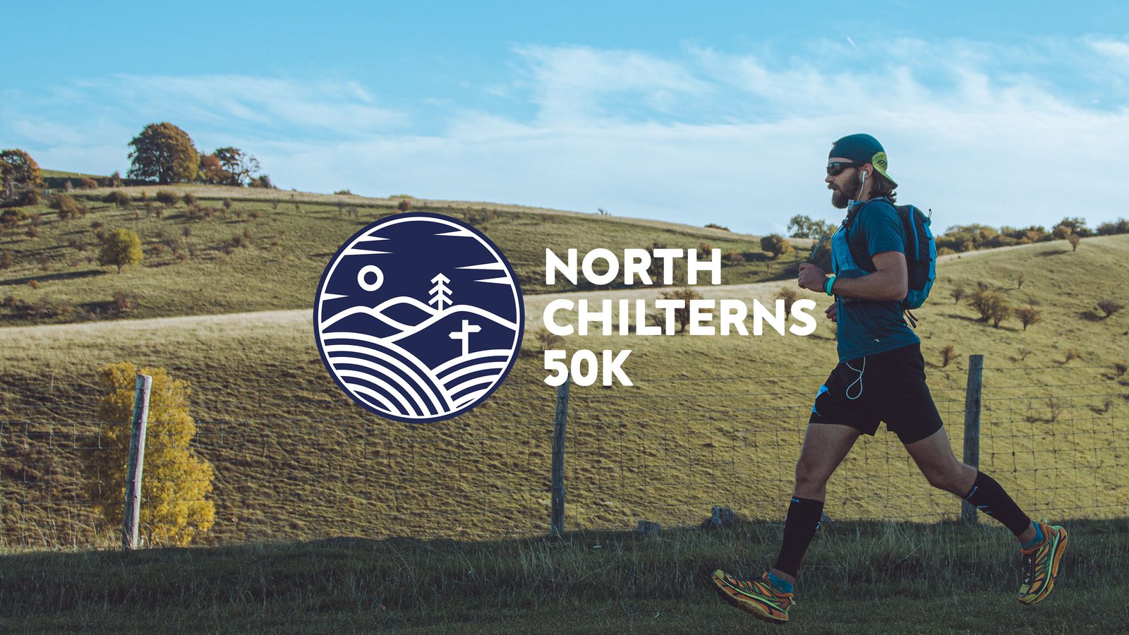 A runner with a blue hydration pack runs along a grassy hillside under a clear sky. The logo for the North Chilterns 50K event is prominently displayed on the left, featuring stylized hills, a tree, a sun or moon, and a cross.
