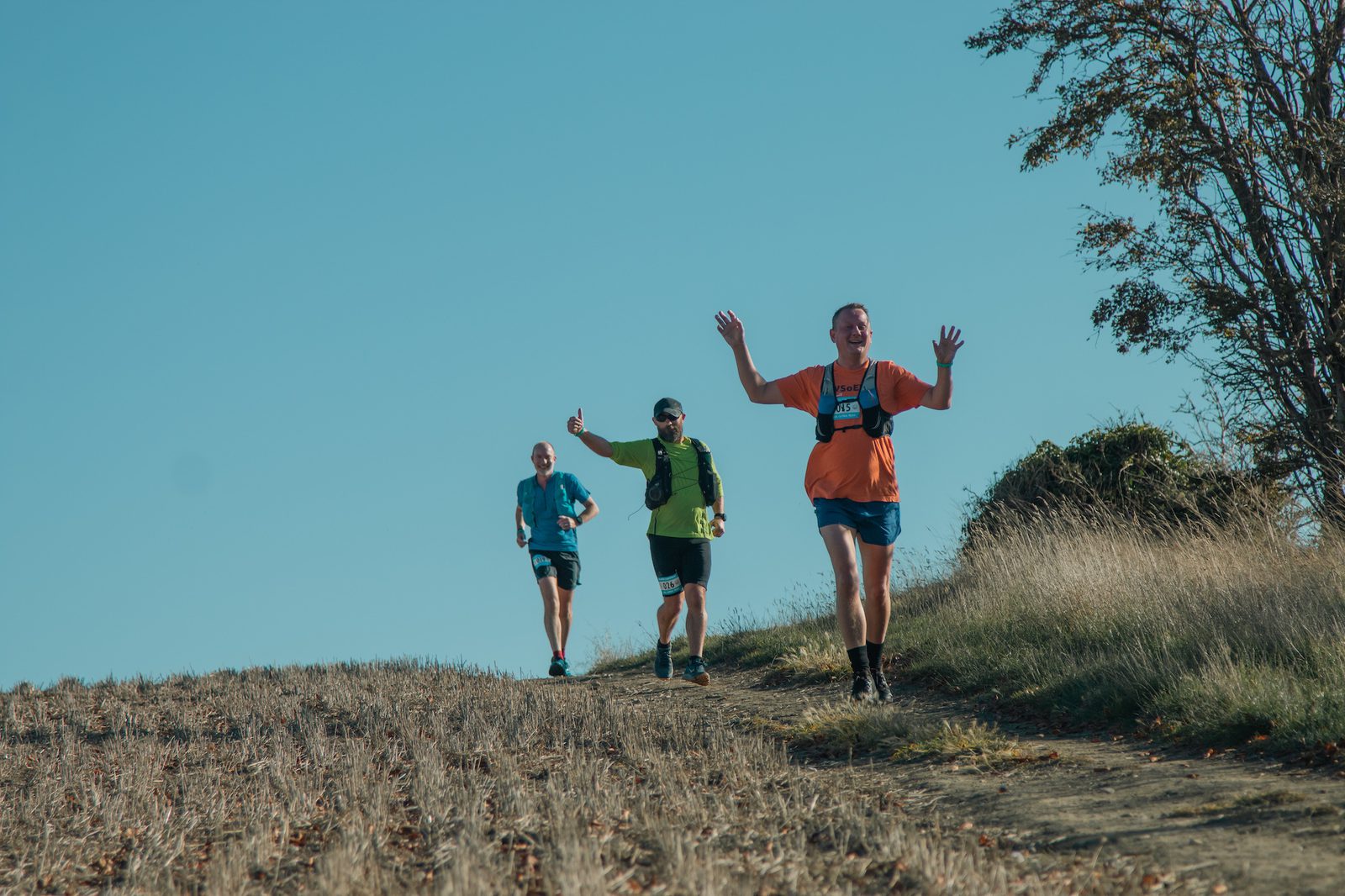 Three runners are participating in a race on a grassy and slightly rocky trail under a clear blue sky. The runner in the foreground is smiling and raising their arms, while the other two runners are closely following behind through the landscape.